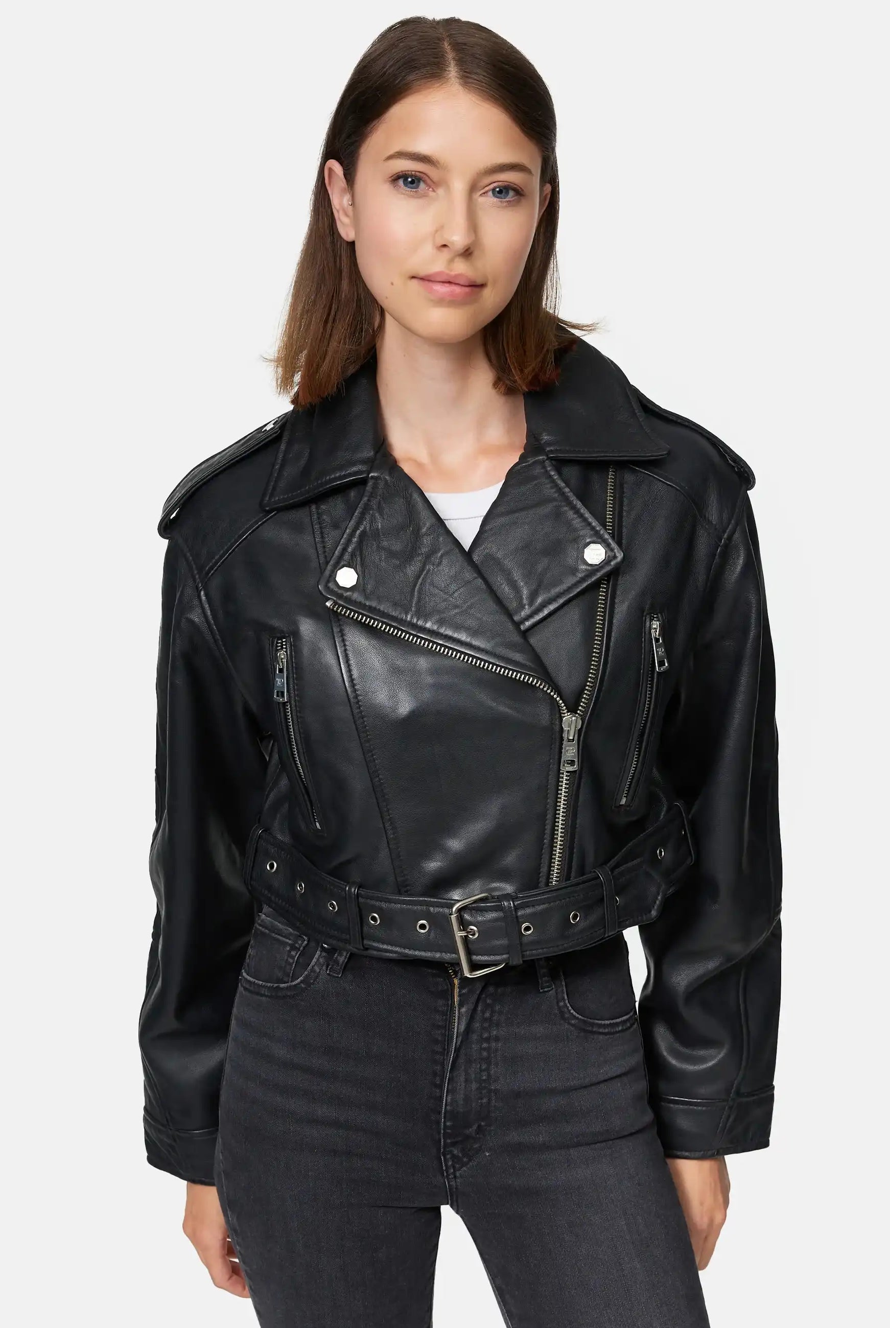 Eine Frau mit glattem braunem Haar trägt eine Trueprodigy Rosa Kurze Bomber-Lederjacke über einem weißen Oberteil und einer schwarzen Jeans mit hoher Taille. Sie steht vor einem schlichten hellen Hintergrund.