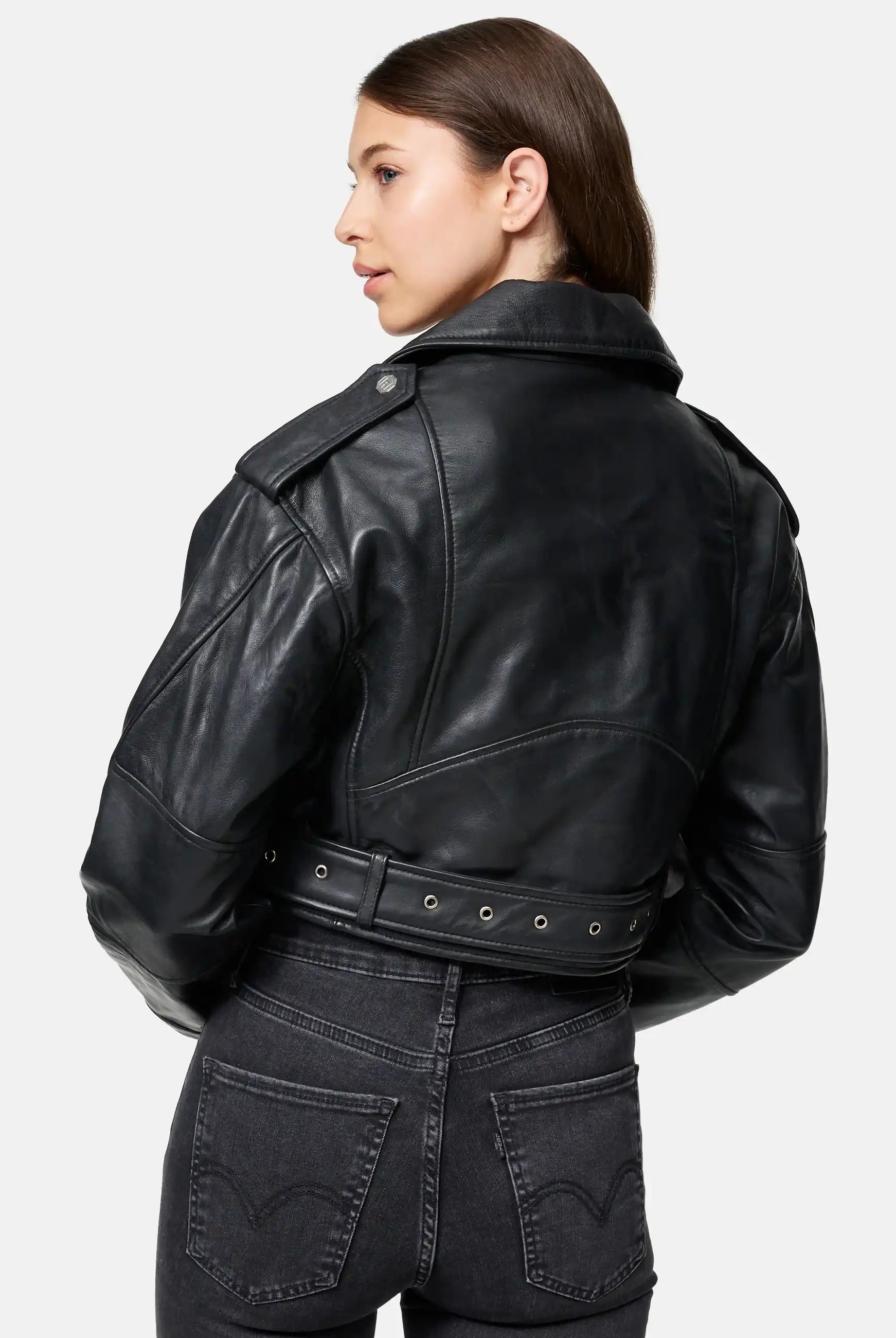 Eine Frau mit langen braunen Haaren trägt die trueprodigy Rosa Kurze Bomber-Lederjacke und schwarze Jeans. Sie steht mit dem Rücken zur Kamera und schaut zur Seite vor einem schlichten hellgrauen Hintergrund.