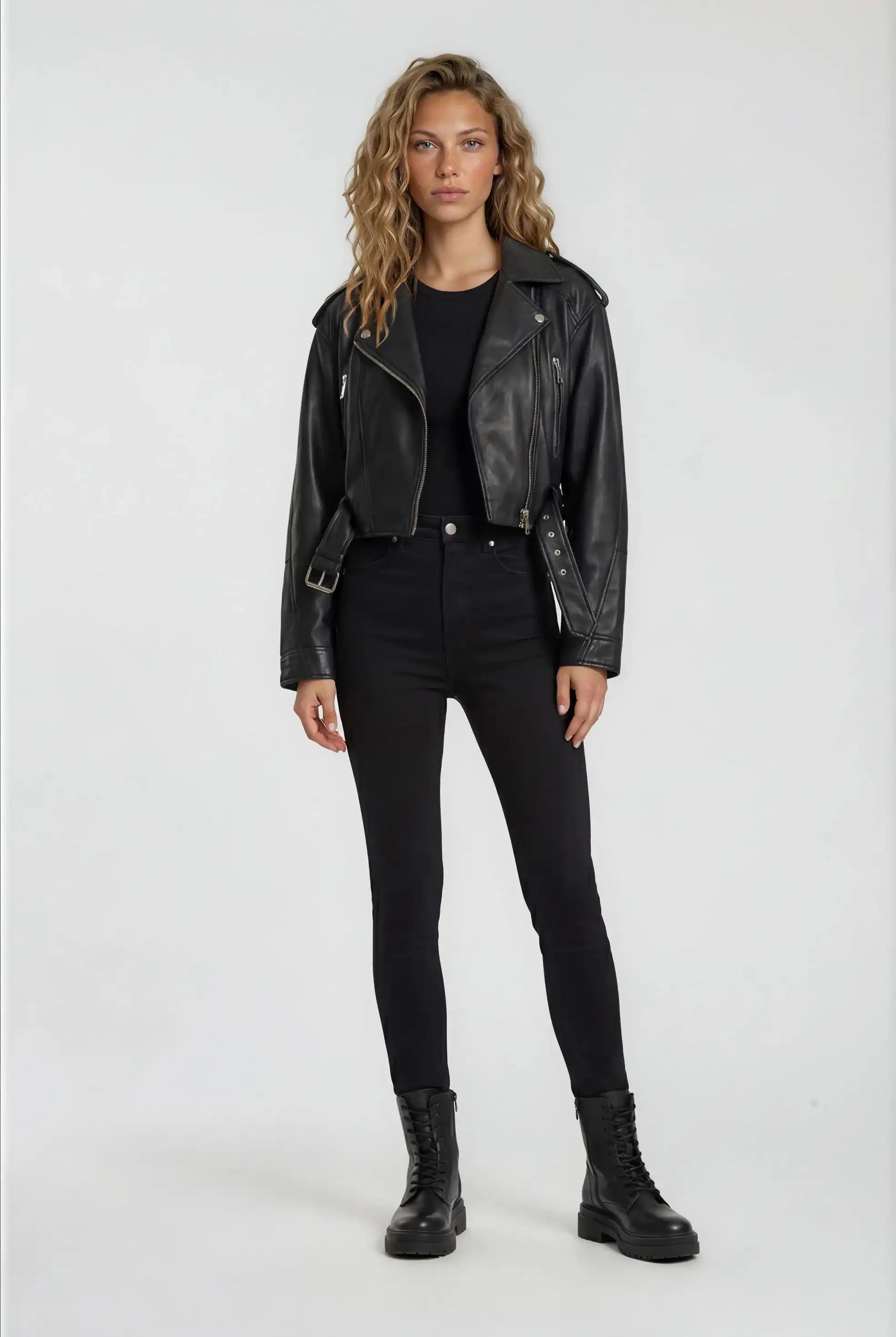 Eine Frau steht mit dem Gesicht nach vorne und trägt eine trueprodigy Kurze Bomber-Lederjacke im Biker-Stil, gepaart mit einem schwarzen T-Shirt, einer schwarzen Hose mit hoher Taille und schwarzen Schnürstiefeln vor einem schlichten hellen Hintergrund.