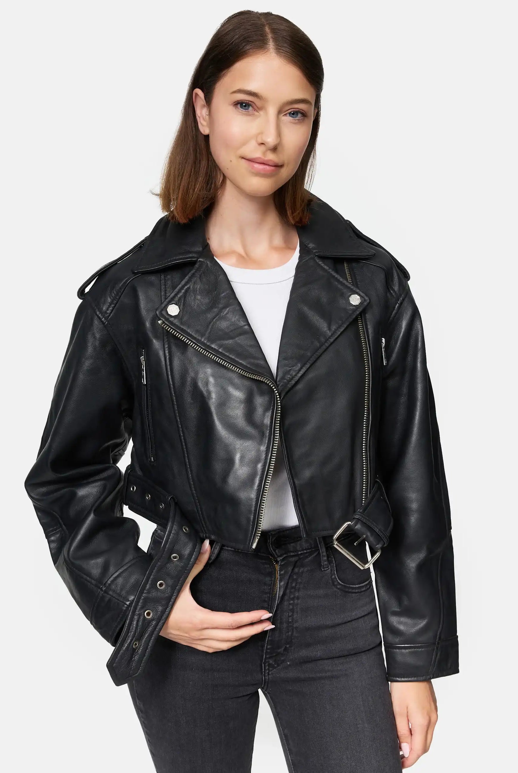 Eine Frau mit glattem braunem Haar trägt die Rosa Kurze Bomber-Lederjacke von trueprodigy über einem weißen Hemd und schwarzen Jeans. Sie steht vor einem schlichten weißen Hintergrund, eine Hand in der Tasche, die andere hält ihre Jacke.