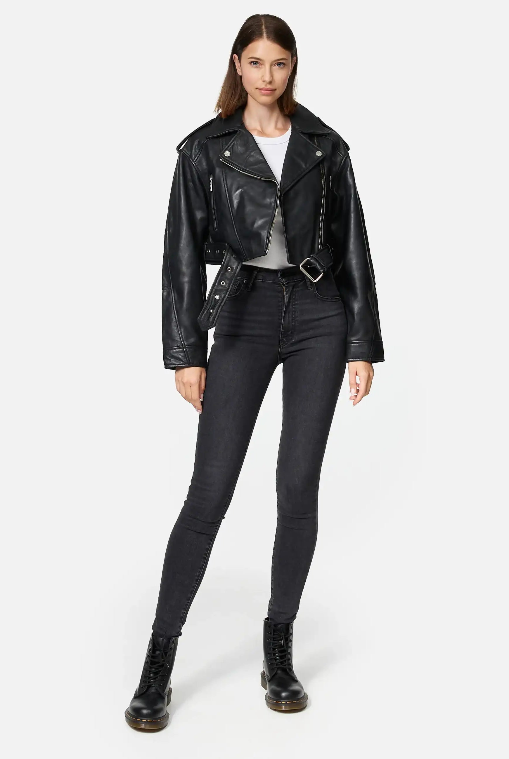 Eine Frau mit glattem, schulterlangem braunem Haar posiert vor einem unifarbenen Hintergrund und trägt eine Rosa Kurze Bomber-Lederjacke von trueprodigy, ein weißes T-Shirt, schwarze Skinny Jeans mit hoher Taille und schwarze Schnürstiefel.