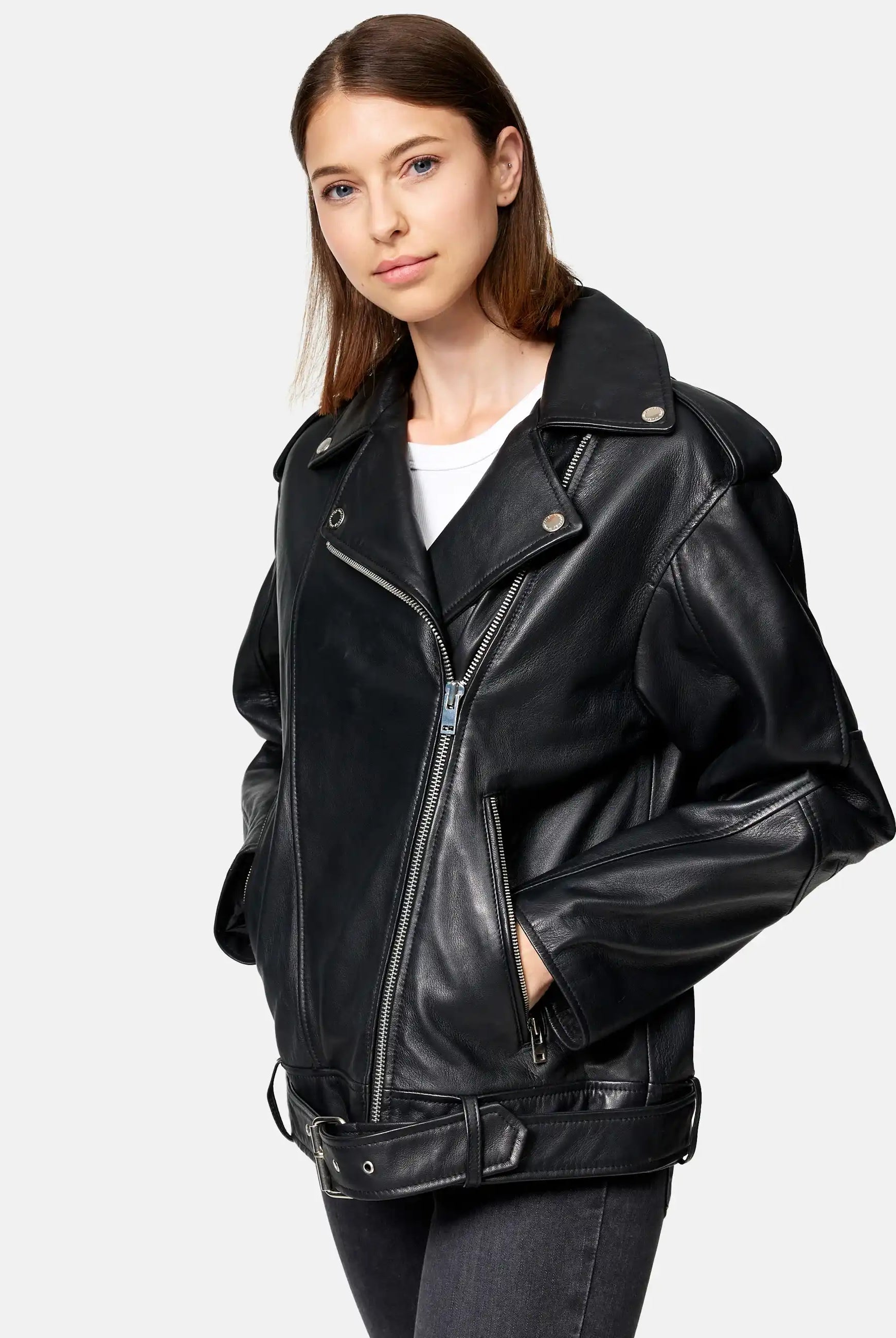 Eine Frau trägt eine trueprodigy Dua Lange übergroße Bikerlederjacke in Schwarz über einem weißen Oberteil und dunklen Jeans und steht mit den Händen in den Jackentaschen vor einem schlichten hellgrauen Hintergrund.