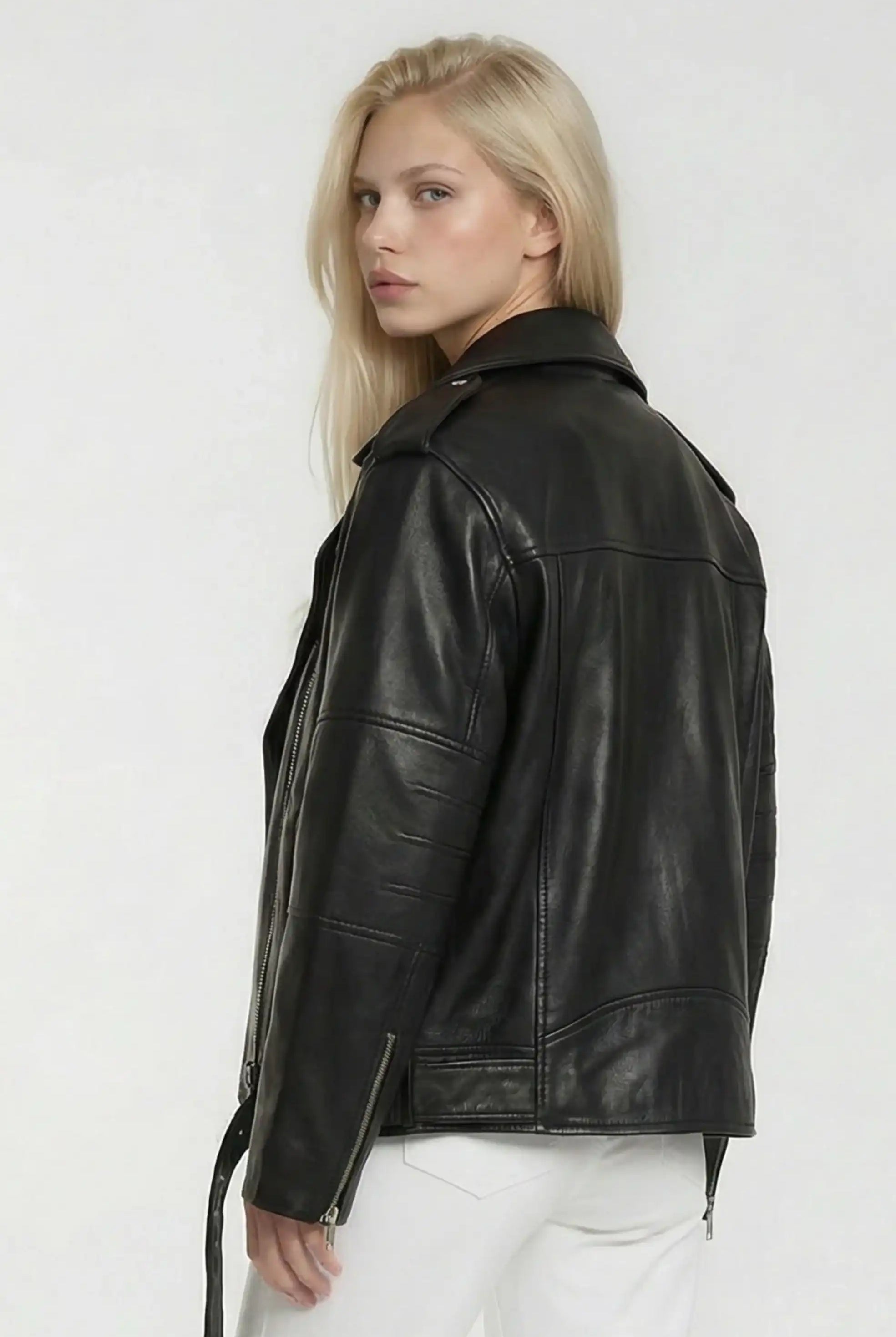 Eine Person mit langen blonden Haaren modelliert die trueprodigy Lange übergroße Bikerlederjacke und weiße Hose, steht vor einem schlichten hellen Hintergrund und schaut über die Schulter in Richtung Kamera.