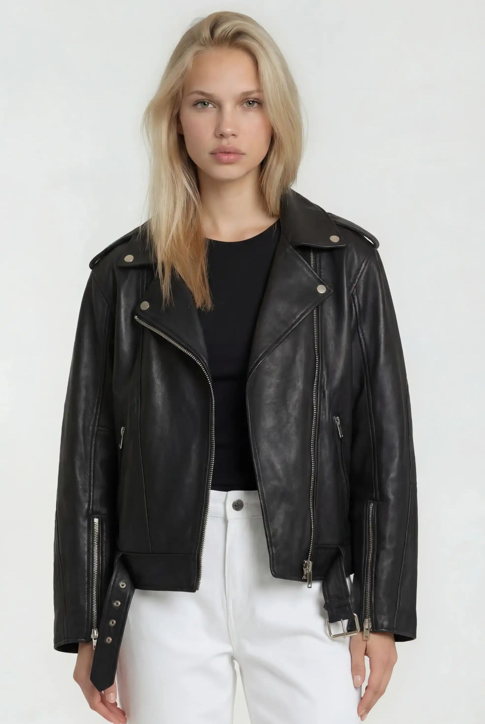 Eine Frau mit langen blonden Haaren trägt eine Lange übergroße Bikerlederjacke von trueprodigy aus weichem Schafsleder, steht vor einem schlichten hellen Hintergrund und schaut mit neutralem Blick direkt in die Kamera.