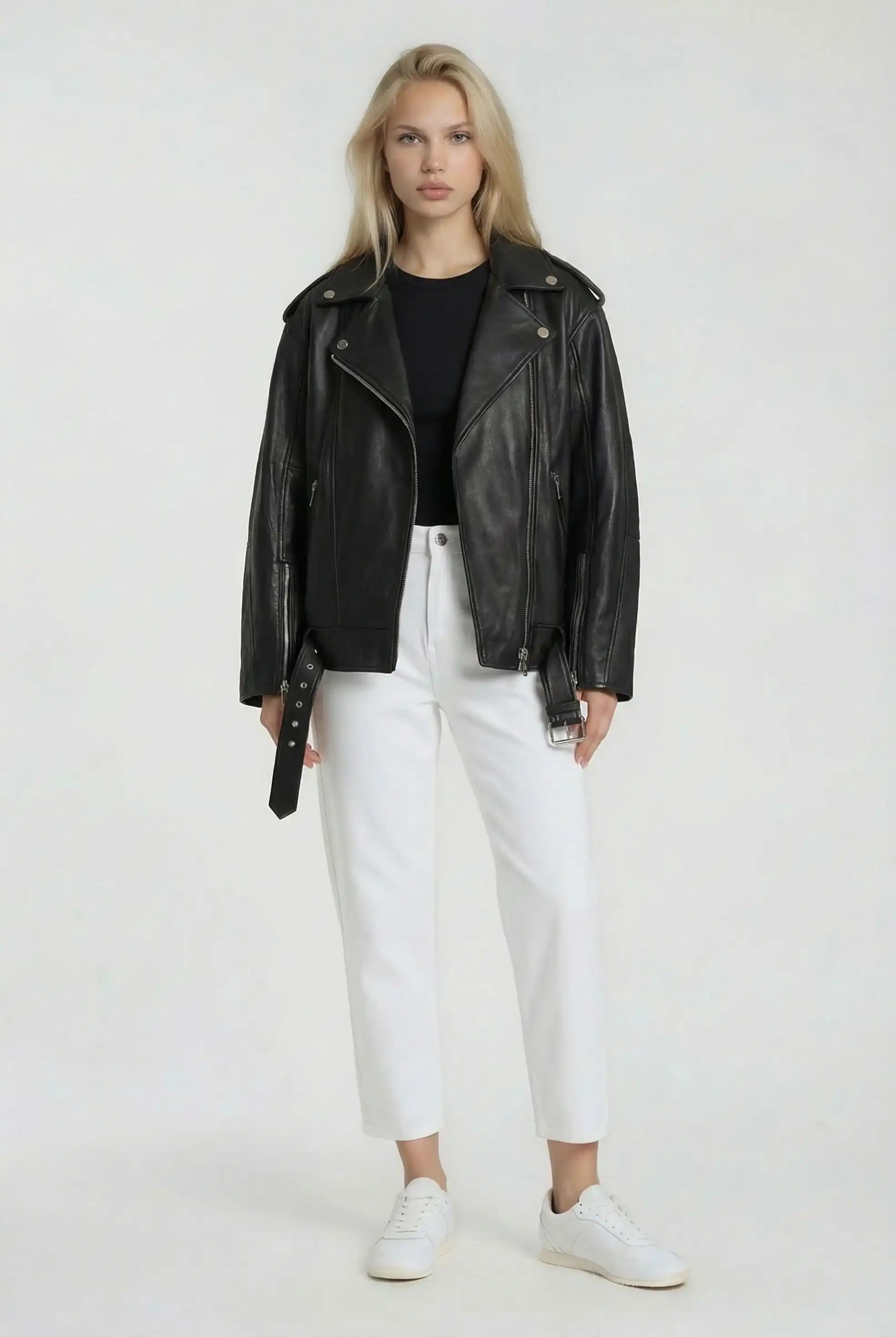 Eine Person steht vor der Kamera und trägt eine schwarze Lange übergroße Bikerlederjacke von trueprodigy, gestylt mit einem schwarzen Hemd, einer weißen Hose mit hoher Taille und weißen Turnschuhen vor einem schlichten hellen Hintergrund.