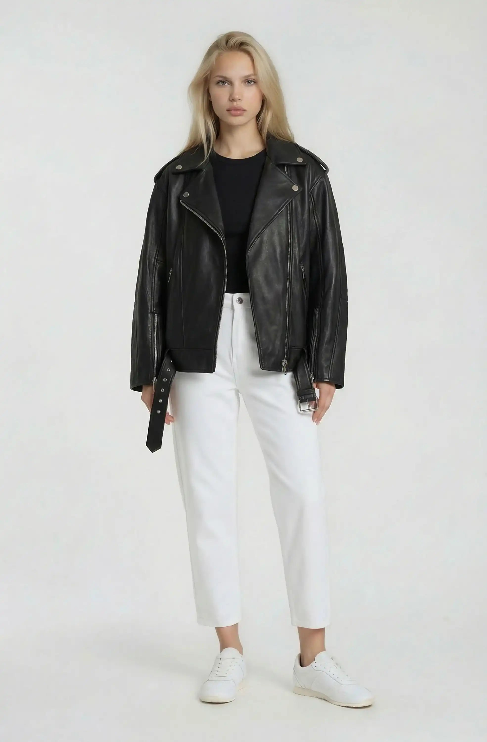 Eine Person steht vor der Kamera und trägt eine schwarze Lange übergroße Bikerlederjacke von trueprodigy, gestylt mit einem schwarzen Hemd, einer weißen Hose mit hoher Taille und weißen Turnschuhen vor einem schlichten hellen Hintergrund.