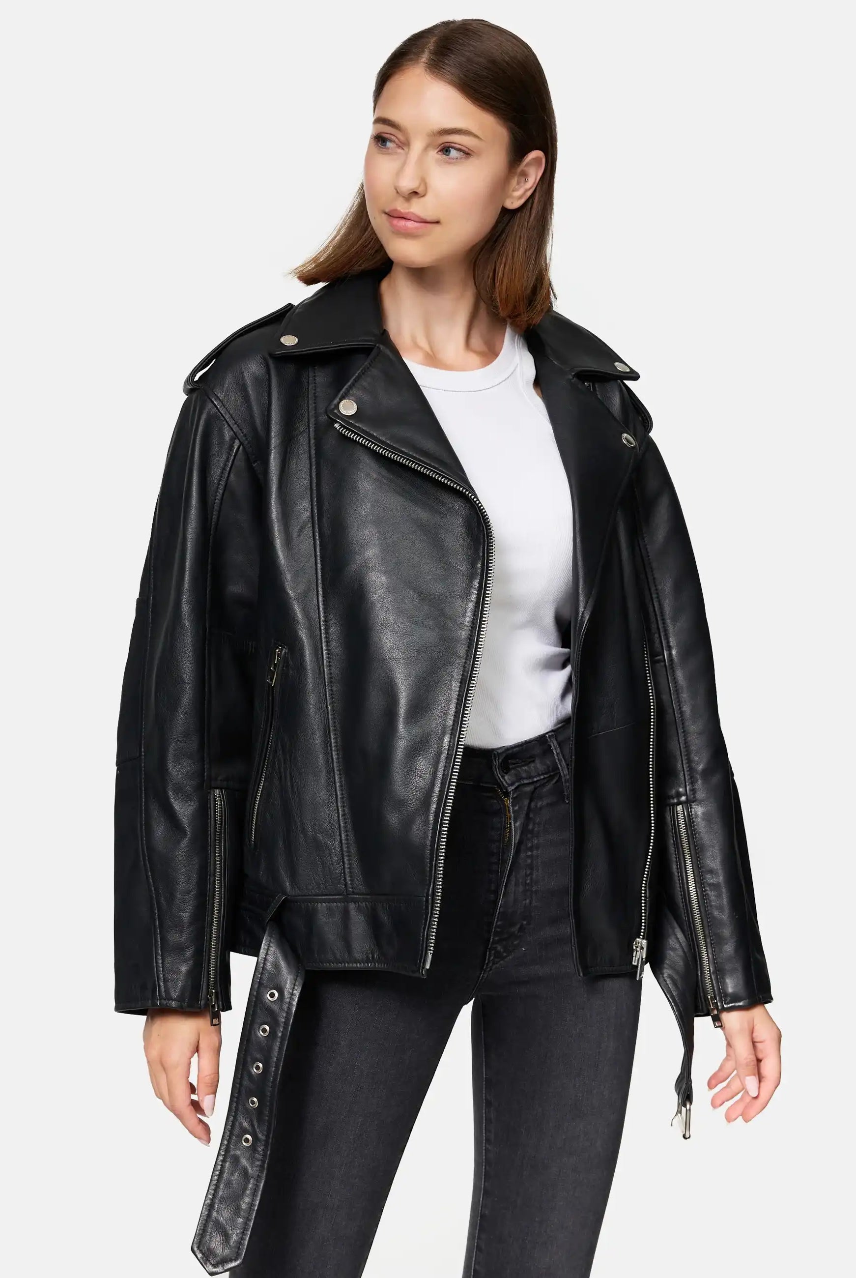 Eine Frau in einer trueprodigy Dua Lange übergroßen Bikerlederjacke, weißem T-Shirt und schwarzen Jeans steht vor einem schlichten hellen Hintergrund und schaut mit neutralem Gesichtsausdruck leicht nach rechts.