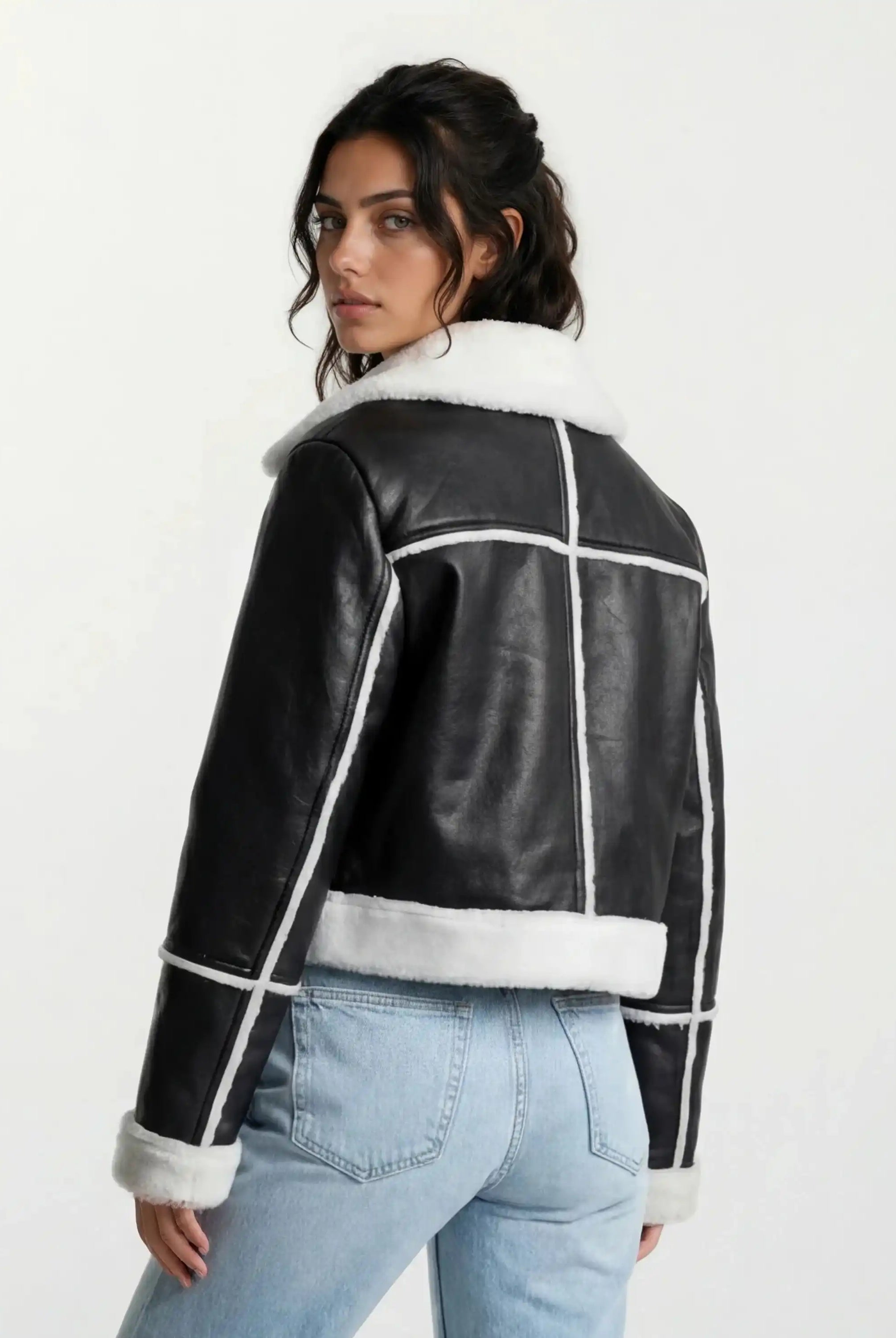 Eine Person mit langen dunklen Haaren trägt eine trueprodigy Damen-Lederjacke mit weißen Teddy-Kontrasten & asymmetrischem Reißverschluss und hellblaue Jeans, steht mit dem Rücken zur Kamera und schaut über die Schulter vor einem schlichten Hintergrund.