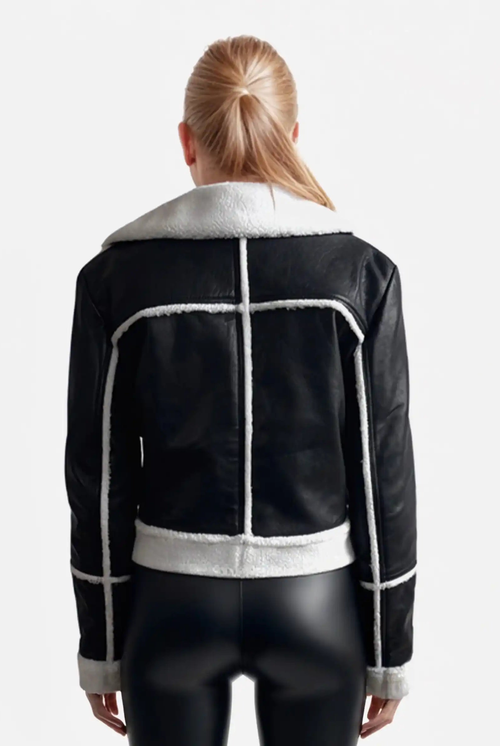Eine Person mit langen blonden Haaren, von hinten gesehen, trägt eine trueprodigy Damen-Lederjacke mit weißen Teddy-Kontrasten & asymmetrischem Zipper in schwarz und taillierter Hose, stehend vor einem schlichten weißen Hintergrund.