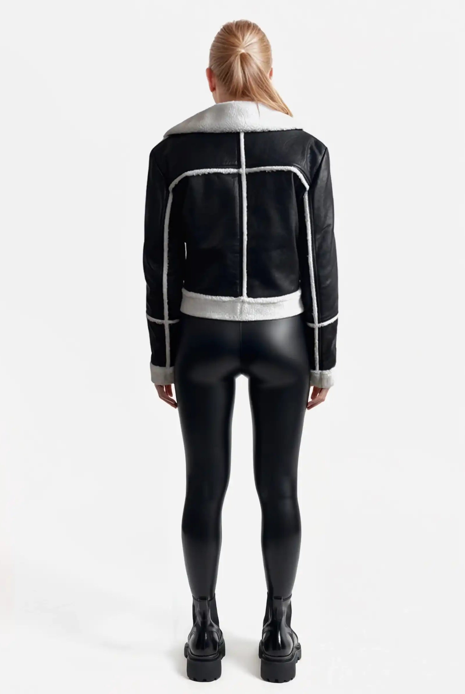 Eine Person mit langen blonden Haaren blickt von sich weg und trägt eine trueprodigy Damen-Lederjacke mit weißen Teddy-Kontrasten & asymmetrischem Reißverschluss, gepaart mit schwarz glänzenden Leggings und schwarzen Plateaustiefeln vor einem schlichten weißen Hintergrund.