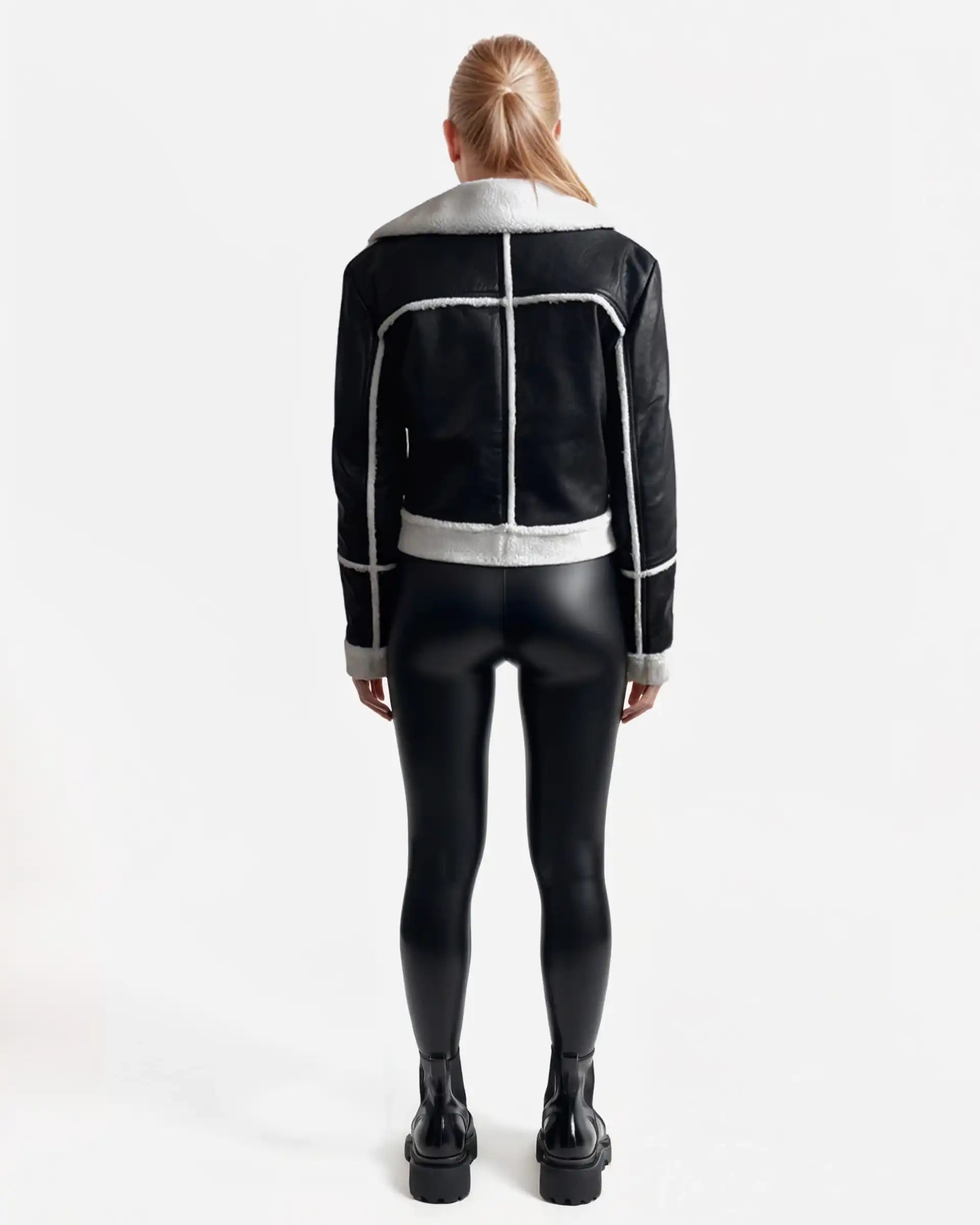 Eine Person mit langen blonden Haaren blickt von sich weg und trägt eine trueprodigy Damen-Lederjacke mit weißen Teddy-Kontrasten & asymmetrischem Reißverschluss, gepaart mit schwarz glänzenden Leggings und schwarzen Plateaustiefeln vor einem schlichten weißen Hintergrund.