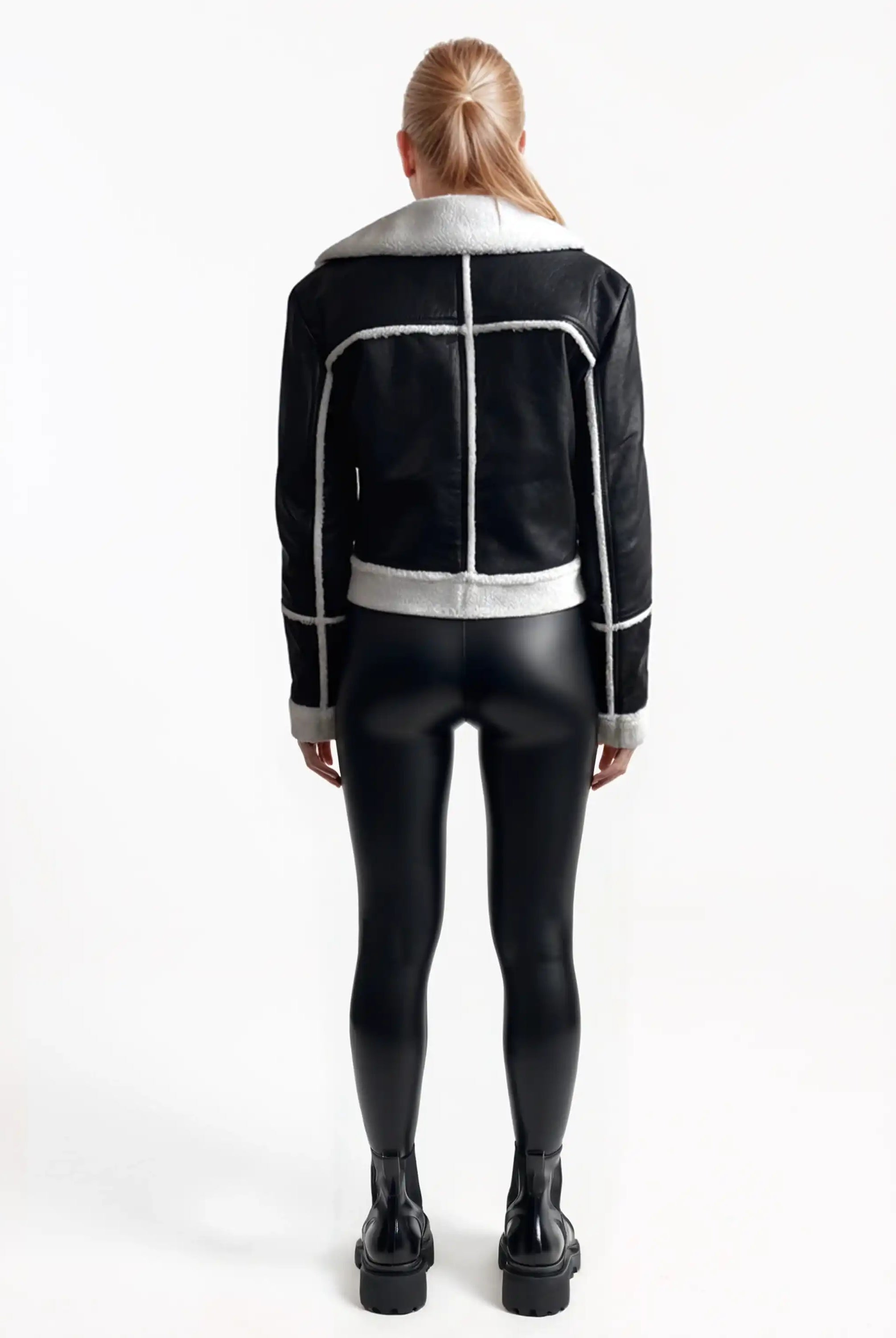 Eine Frau mit langem, glattem blondem Haar blickt nach vorne und trägt die trueprodigy Damen-Lederjacke mit weißen Teddy-Kontrasten & asymmetrischem Reißverschluss, gepaart mit schwarz glänzenden Leggings und Stiefeletten vor einem schlichten weißen Hintergrund.