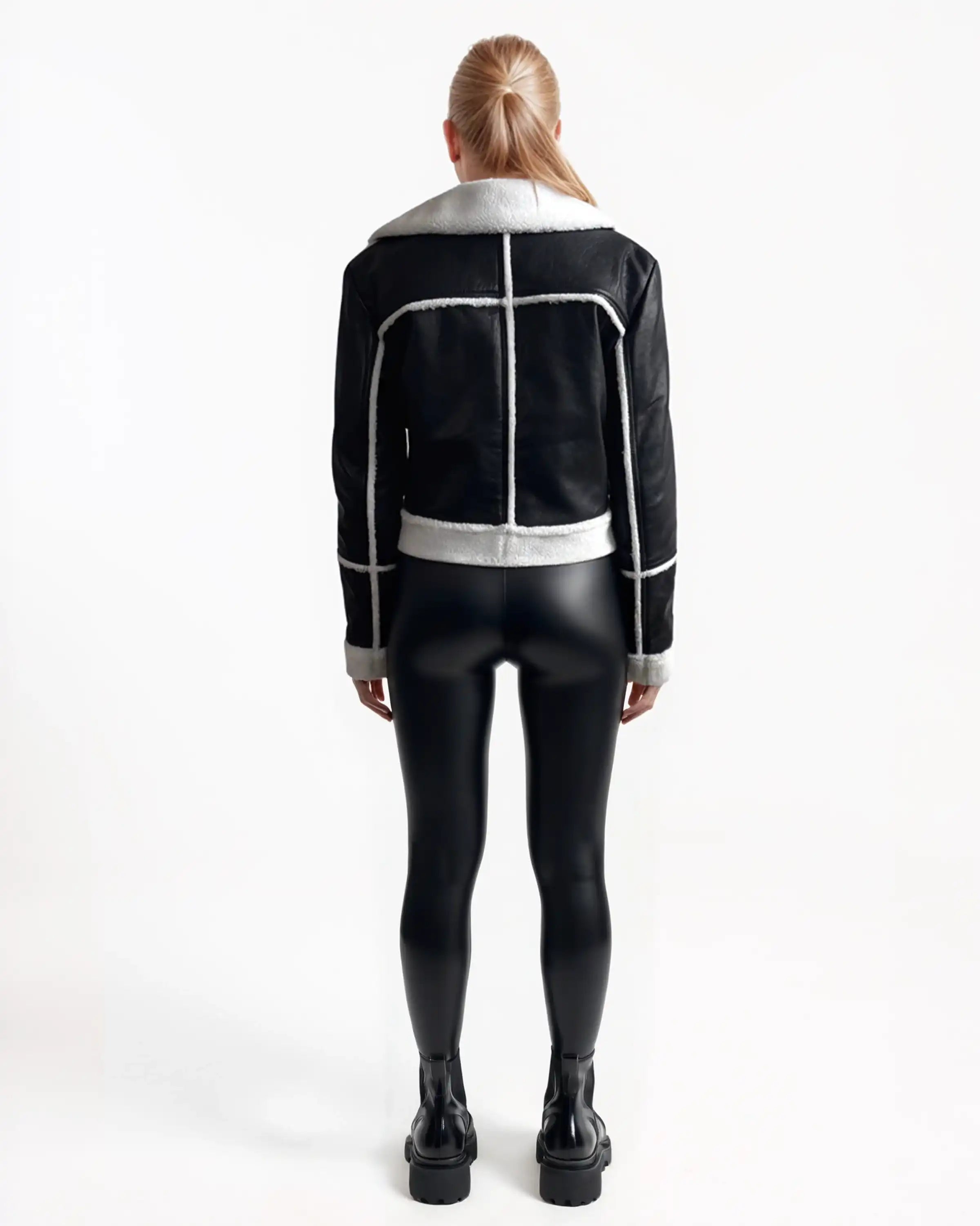 Eine Frau mit langem, glattem blondem Haar blickt nach vorne und trägt die trueprodigy Damen-Lederjacke mit weißen Teddy-Kontrasten & asymmetrischem Reißverschluss, gepaart mit schwarz glänzenden Leggings und Stiefeletten vor einem schlichten weißen Hintergrund.