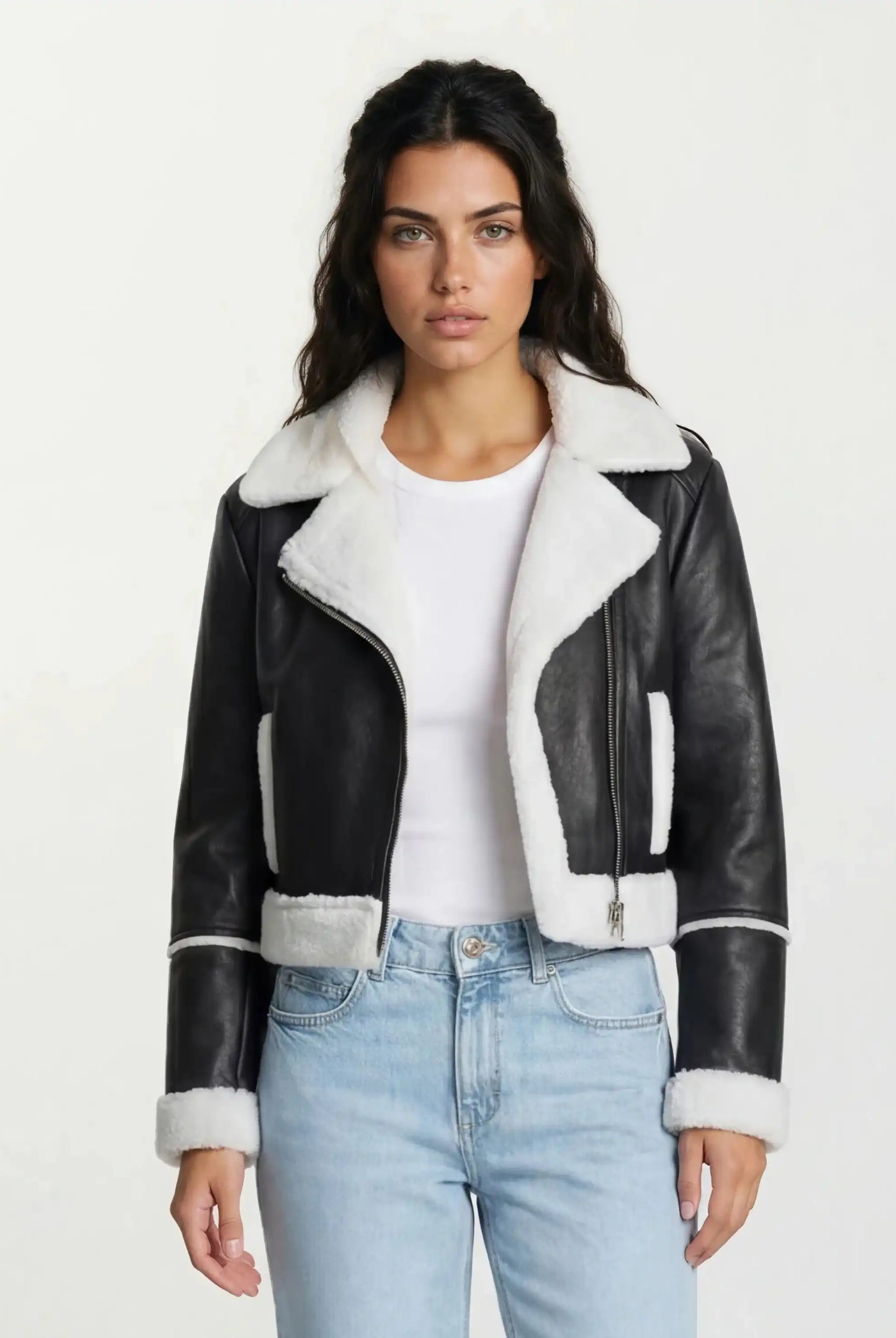 Eine Frau mit langen dunklen Haaren trägt die trueprodigy Damen-Lederjacke mit weißen Teddy-Kontrasten & asymmetrischem Reißverschluss über einem weißen T-Shirt und hellblauen Jeans, vor einem schlichten hellen Hintergrund.