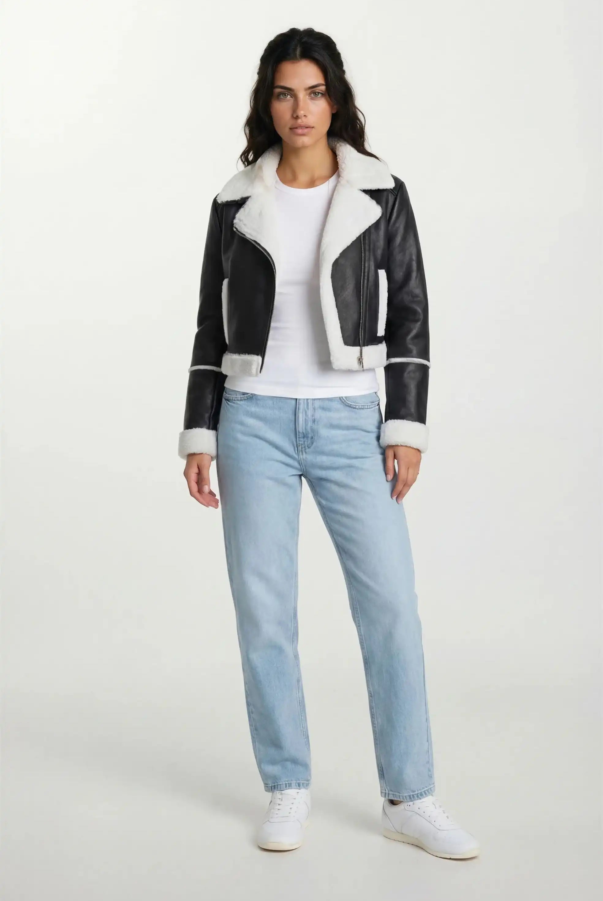 Eine Frau sieht nach vorne in einer trueprodigy Damen-Lederjacke mit weißen Teddy-Kontrasten & asymmetrischem Reißverschluss über einem weißen T-Shirt, hellblauen Jeans und weißen Turnschuhen vor einem schlichten hellen Hintergrund.