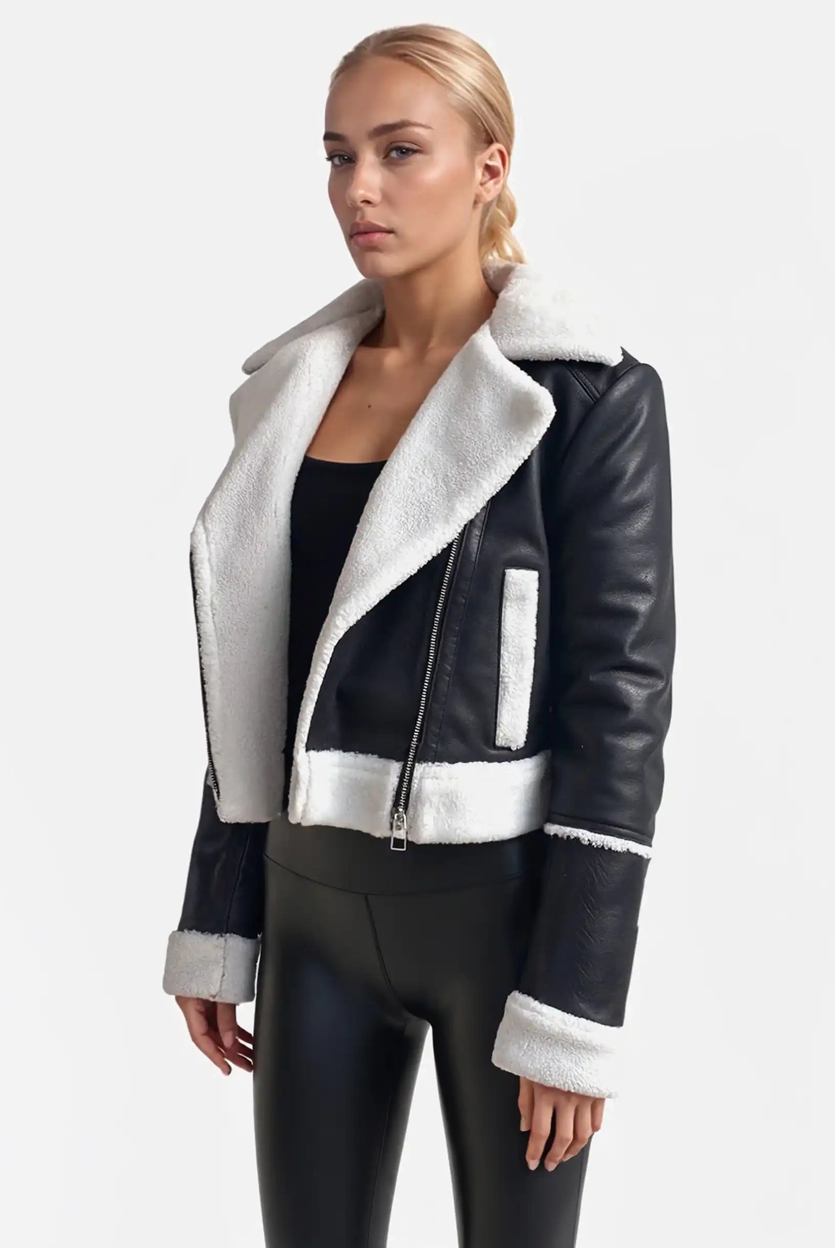 Vor weißem Hintergrund trägt eine Frau von trueprodigy eine Damen-Lederjacke mit weißen Teddy-Kontrasten und asymmetrischem Reißverschluss, dazu ein schwarzes Top und glänzende Hosen. Sie hat blonde, zurückgebundene Haare und blickt neutral.