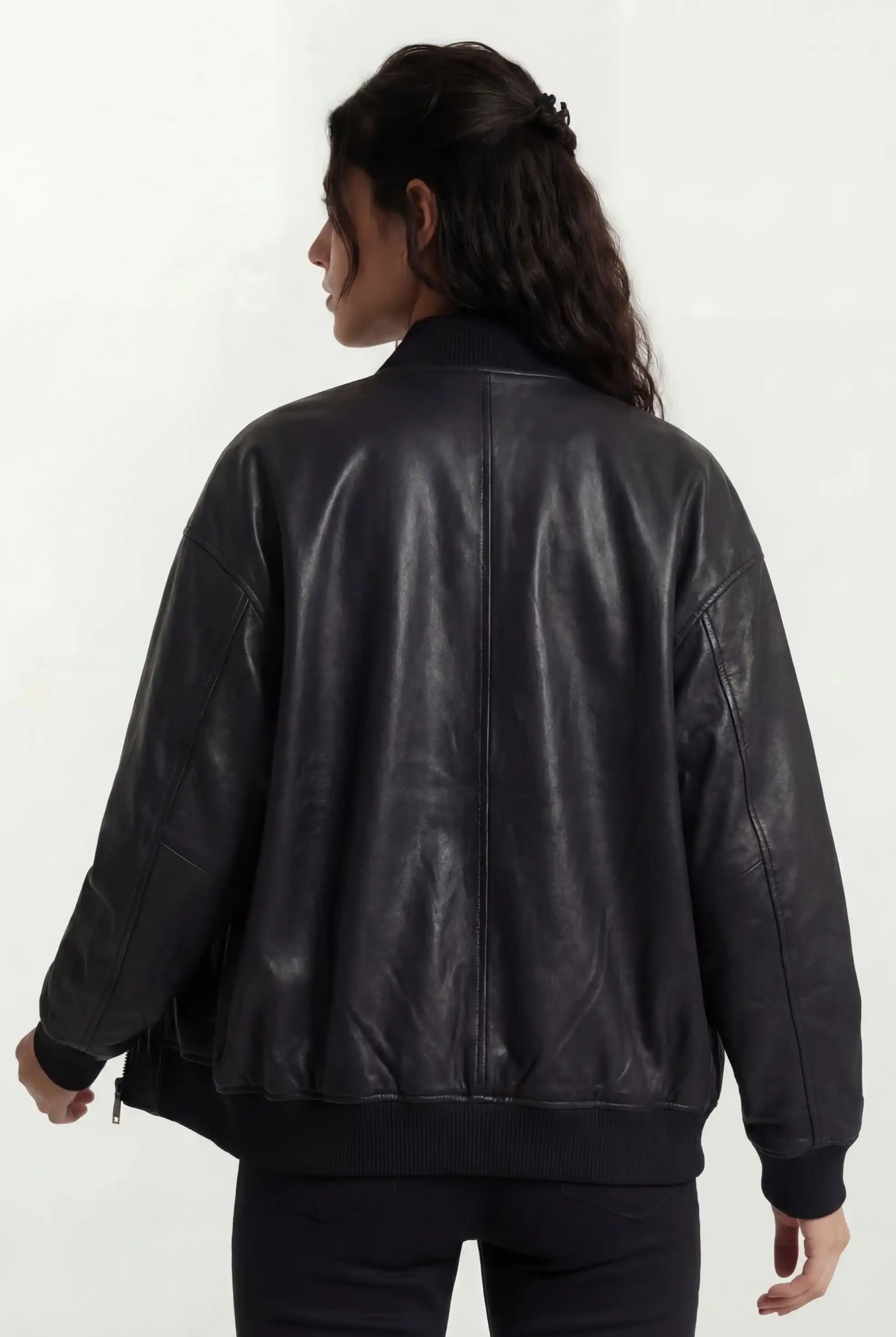 Eine Person mit langen, gewellten dunklen Haaren wendet sich von der Kamera ab und trägt eine trueprodigy Oversize-Bomberjacke aus Leder mit Rippbündchen & schwarzem Zipper und eine dunkle Hose vor einem schlichten hellen Hintergrund.