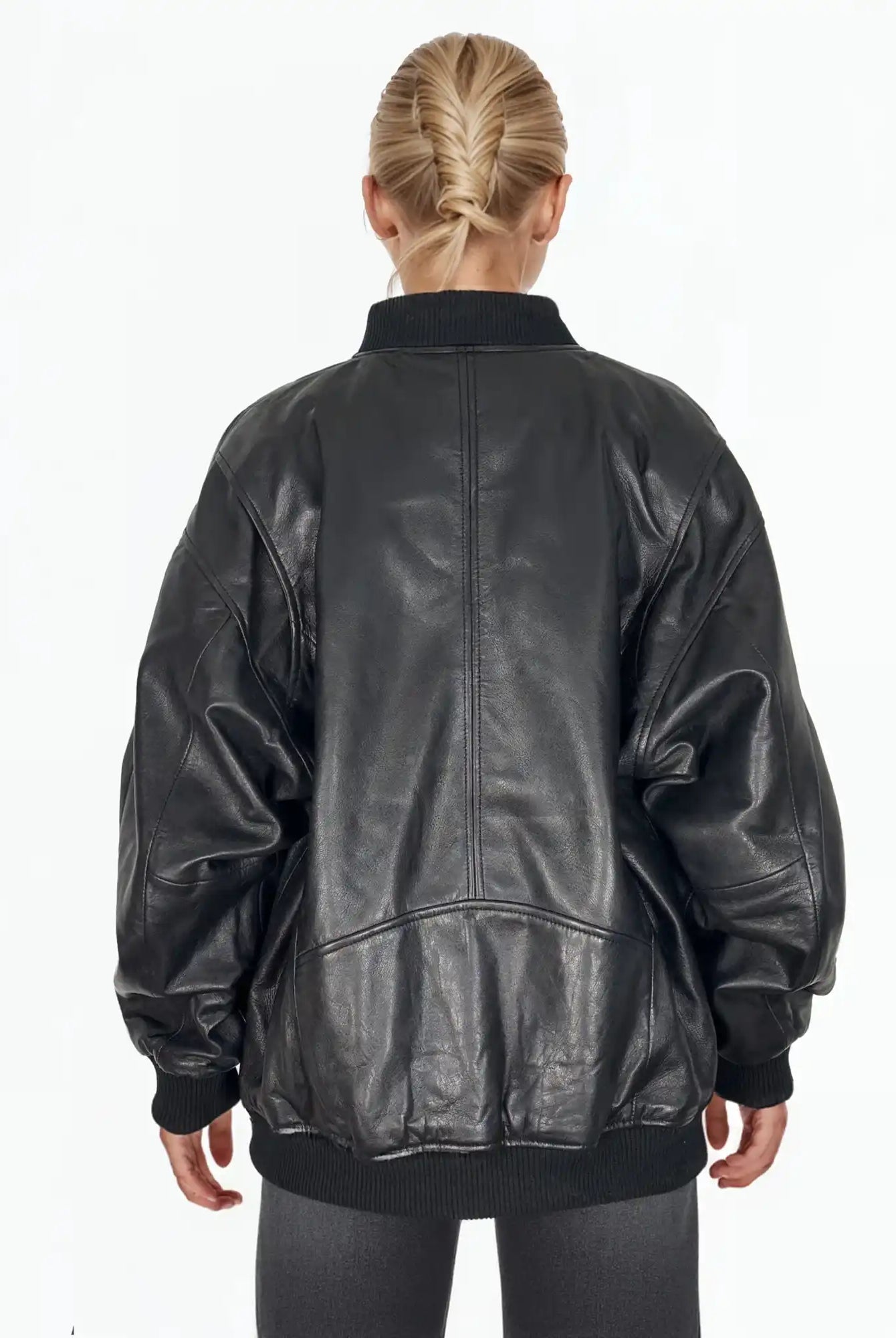 Eine Person mit blondem Haar in einem tiefen Zopf ist von hinten zu sehen. Sie trägt die trueprodigy Selina Oversize-Bomberjacke aus Leder mit Rippbündchen & schwarzem Zipper, gepaart mit einer dunklen Hose vor einem schlichten weißen Hintergrund.