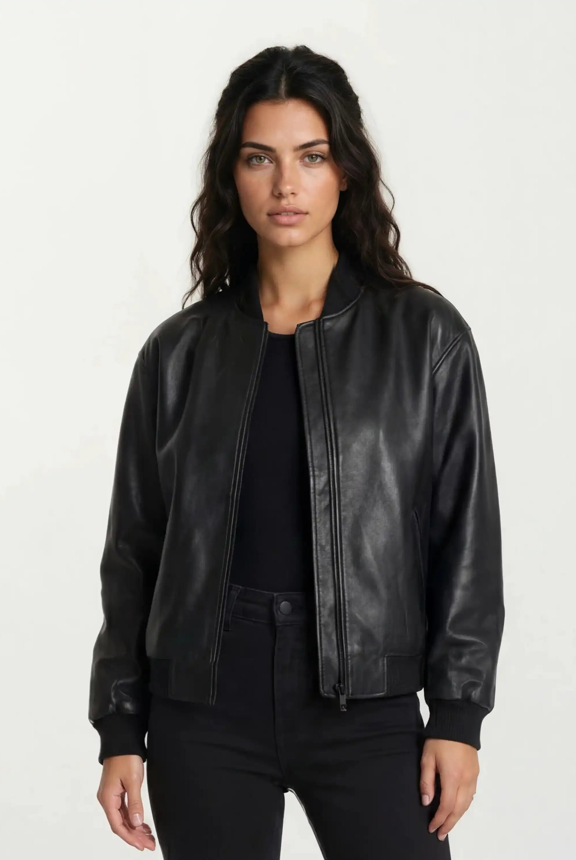 Eine Frau mit langen, gewellten dunklen Haaren trägt eine trueprodigy Oversize-Bomberjacke aus Leder mit Rippbündchen & schwarzem Reißverschluss über einem schwarzen Oberteil und einer Hose. Sie steht vor einem schlichten hellen Hintergrund und blickt mit neutralem Gesichtsausdruck nach vorne.