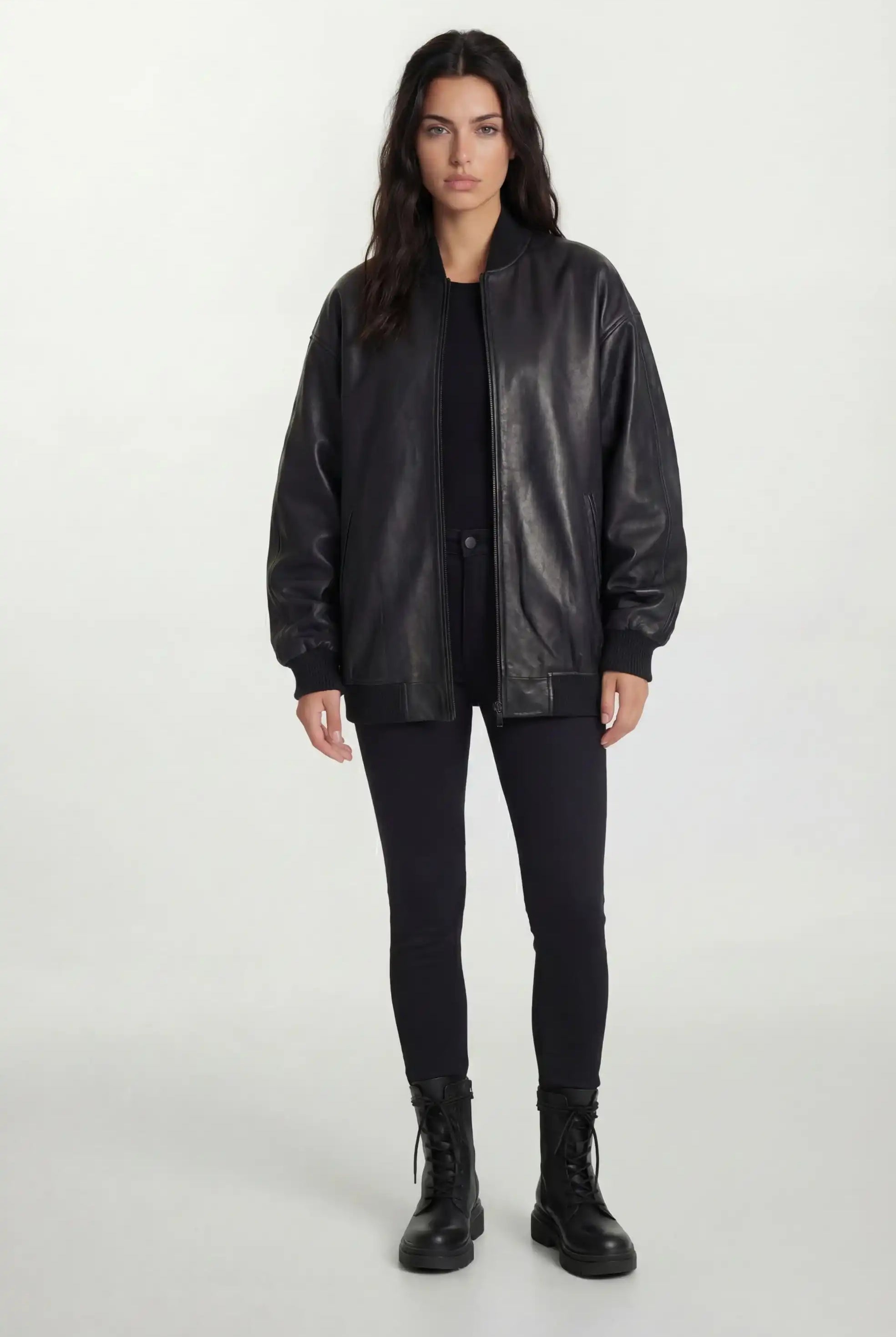 Eine Frau steht vor einem schlichten, hellen Hintergrund und trägt die trueprodigy Oversize-Bomberjacke aus Leder mit Rippbündchen & schwarzem Reißverschluss, gepaart mit einem schwarzen Hemd, Skinny Jeans und Schnürstiefeln, die Arme an den Seiten.