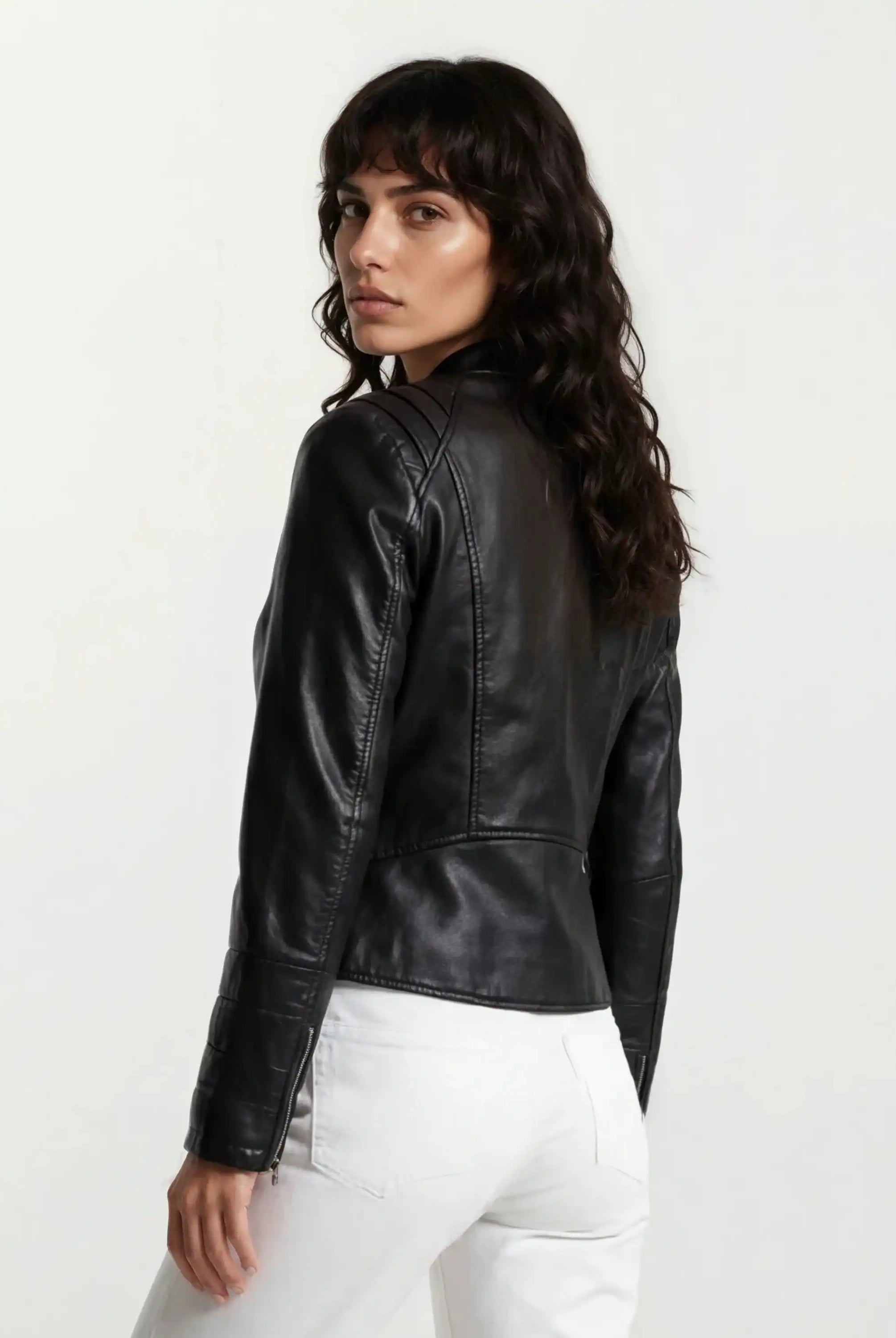 Eine Frau mit langen, gewellten braunen Haaren steht seitlich in weißen Hosen und der trueprodigy Taillierte Lederjacke mit Stehkragen, Steppnähten & silbernen Reißverschlüssen vor einem hellen Hintergrund.