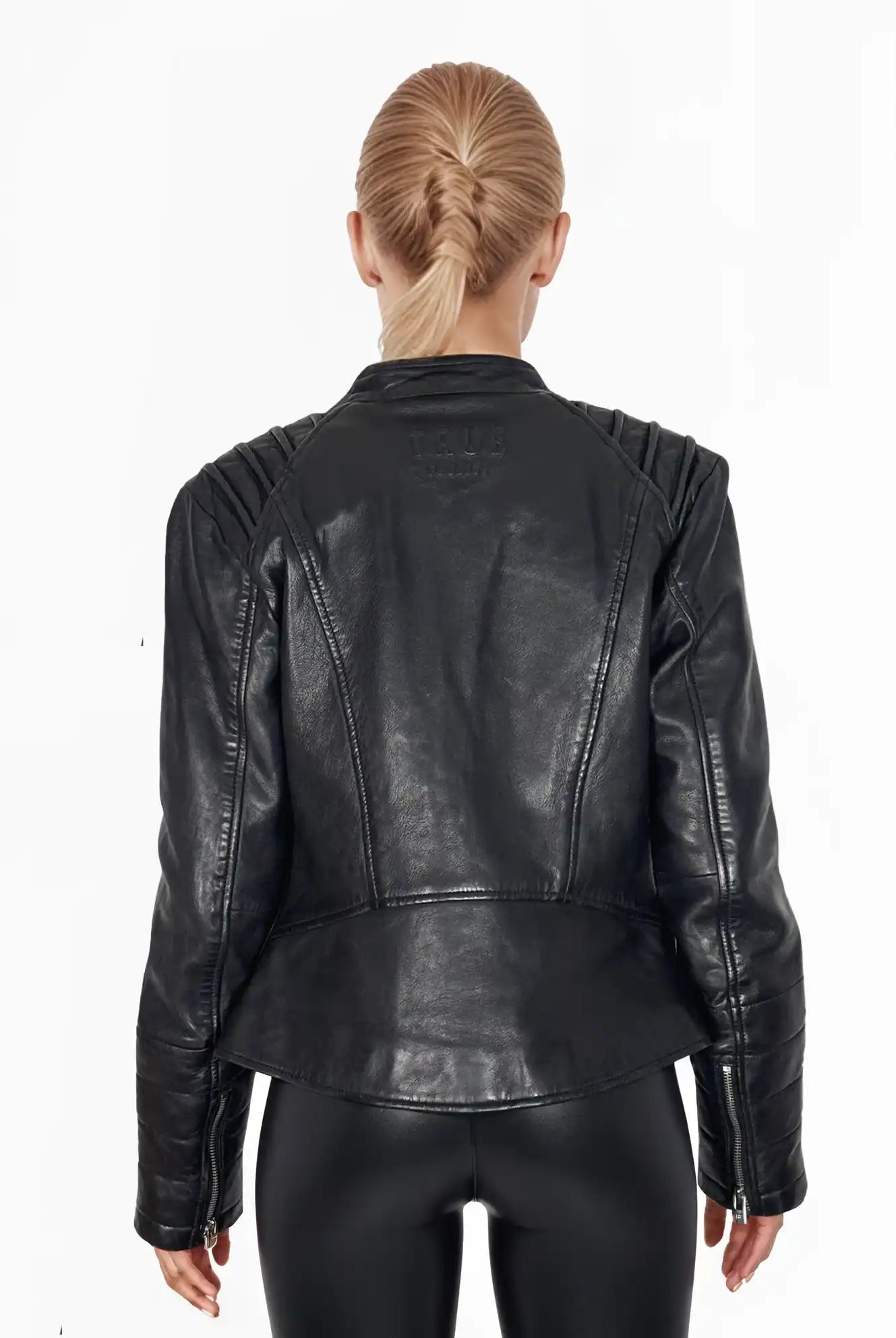 Eine Person mit blonden, geflochtenen Haaren trägt von hinten sichtbar die schwarze Raegan Taillierte Lederjacke mit Stehkragen, Steppnähten & silbernen Reißverschlüssen von trueprodigy sowie eine schwarze Hose vor weißem Hintergrund.