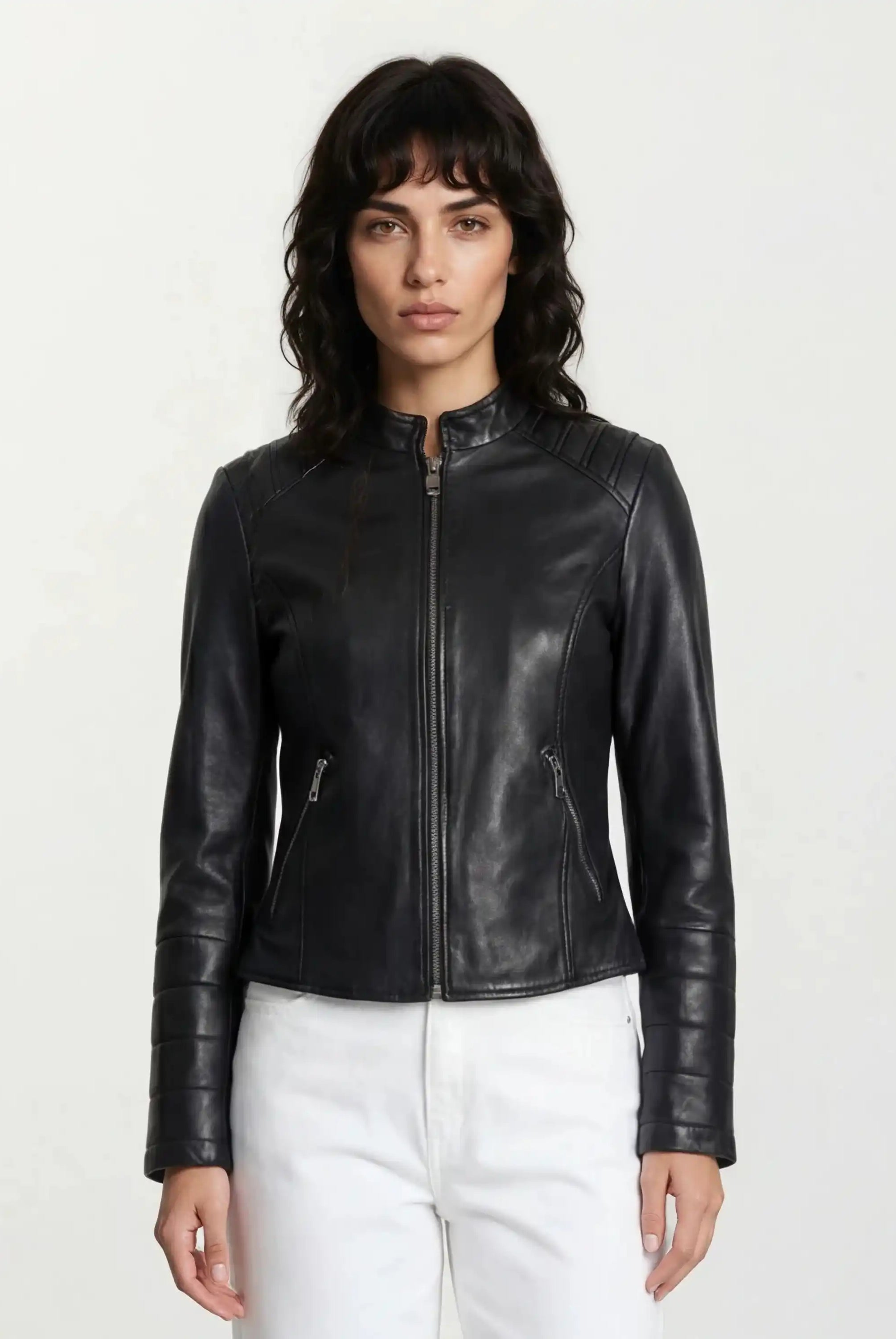 Eine Person mit langen, gewellten dunklen Haaren trägt die taillierte trueprodigy Lederjacke mit Stehkragen, Steppnähten & silbernen Reißverschlüssen und weißen Hosen, steht vor hellem Hintergrund und blickt neutral in die Kamera für einen rockigen Look.