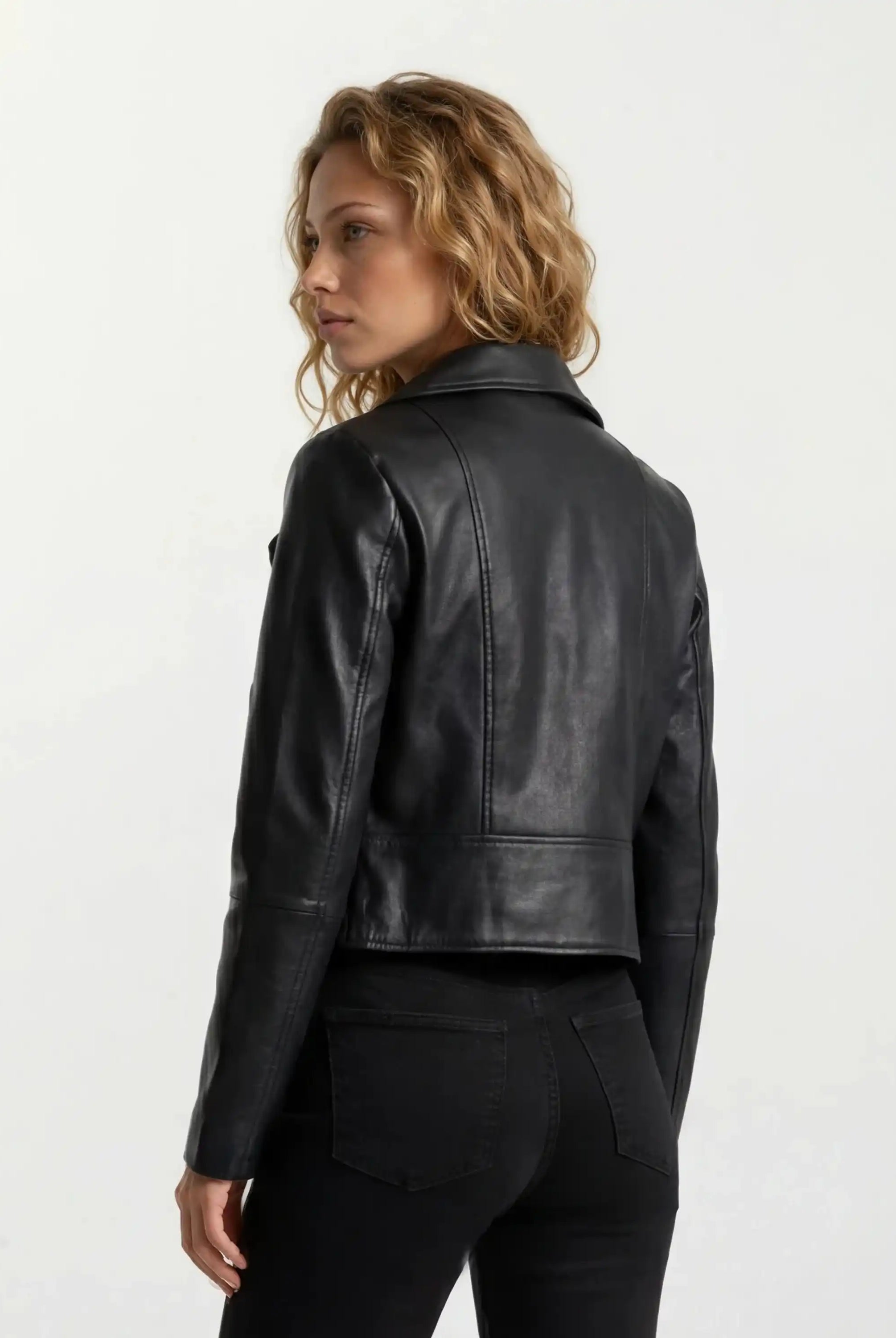 Eine Frau mit gewelltem, schulterlangem blondem Haar wird von hinten und von der Seite gezeigt. Sie trägt eine trueprodigy Taillierte Lederjacke mit schwarzem Reißverschluss & Ärmelzipper und schwarze Jeans vor einem schlichten, hellen Hintergrund.