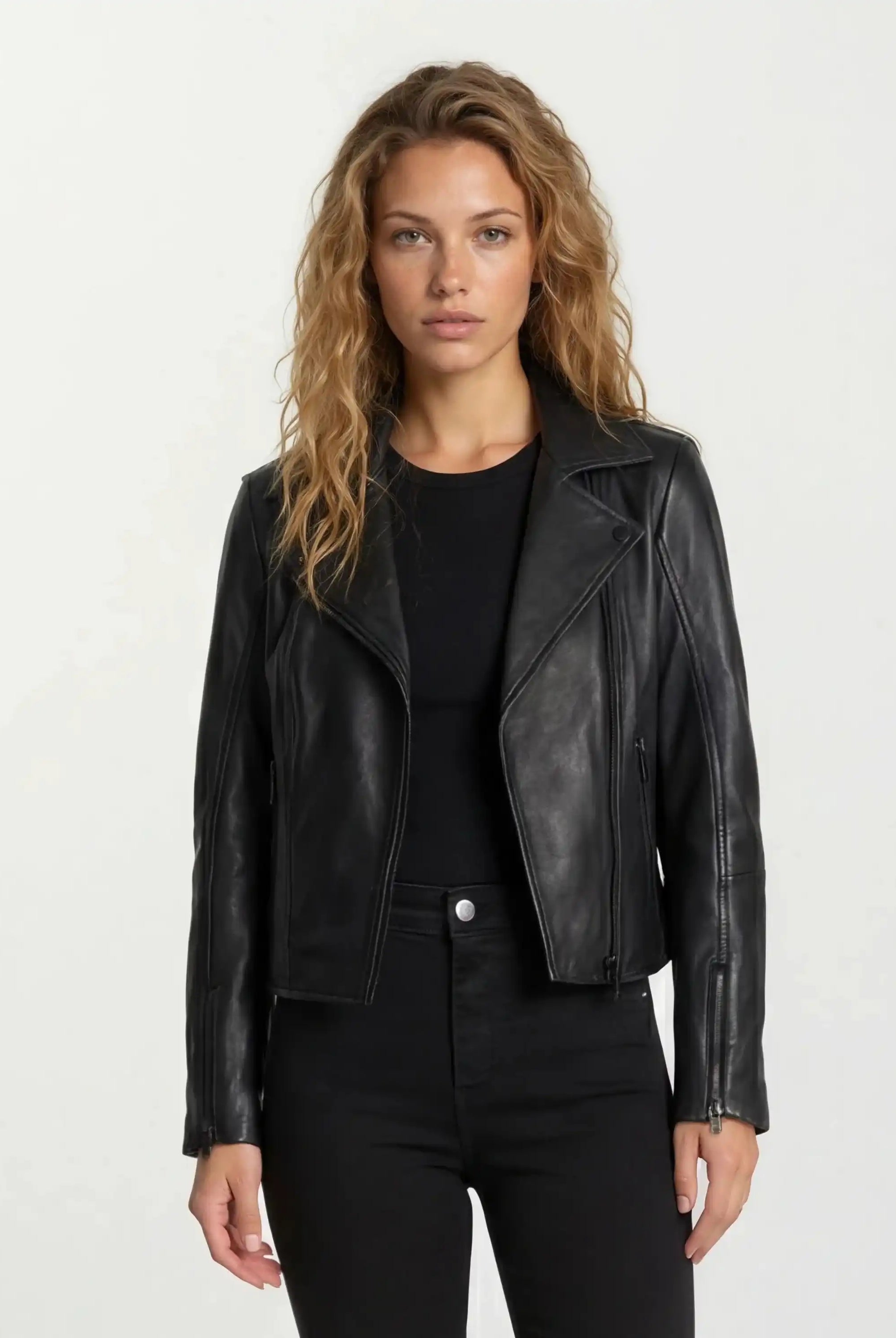 Eine Frau mit langen, welligen blonden Haaren trägt die taillierte Lederjacke von trueprodigy mit schwarzem Reißverschluss und Ärmelzipper über schwarzem Shirt und Hose. Sie steht vor hellem Hintergrund und blickt neutral in die Kamera.