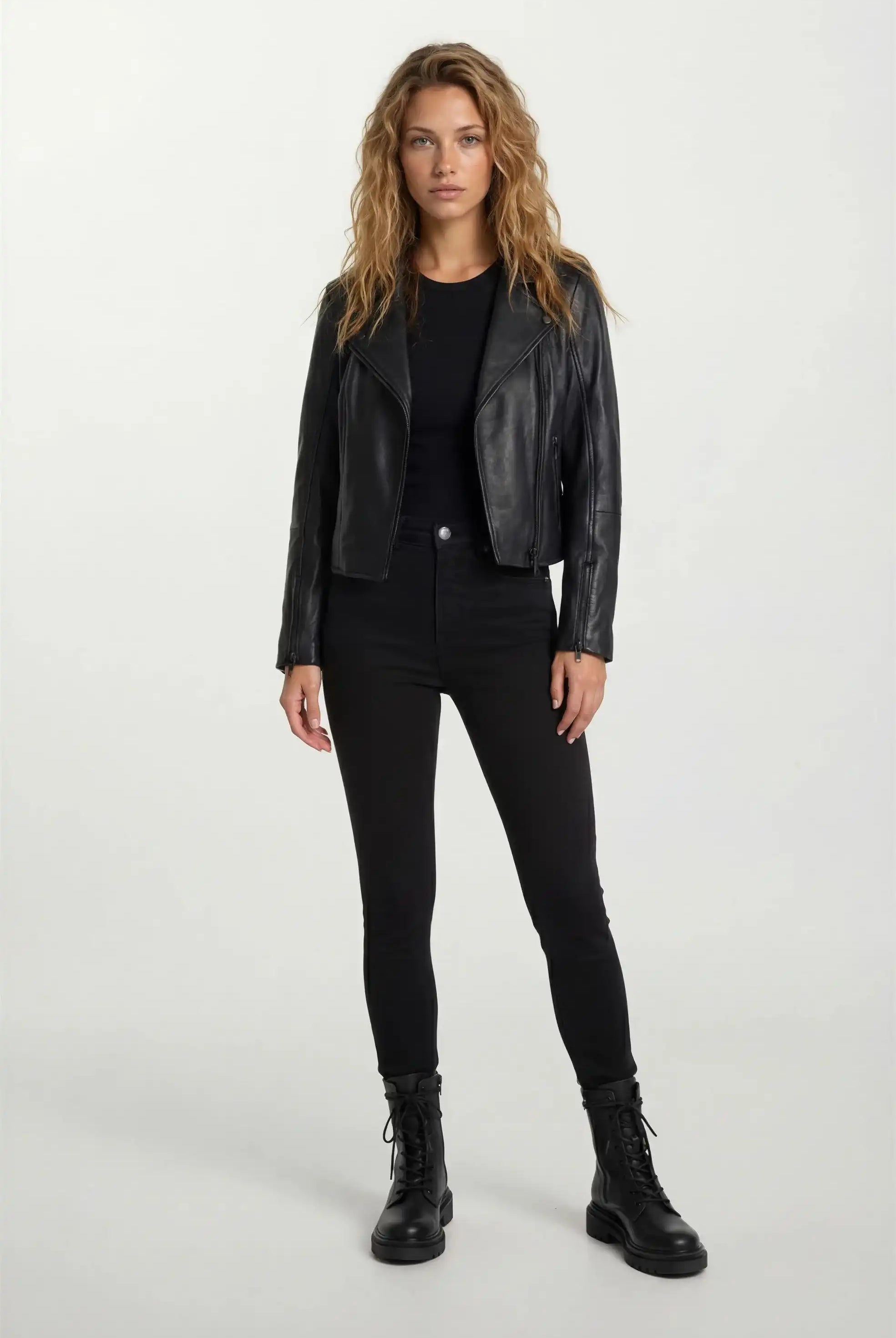 Eine Frau mit langem, gewelltem Haar steht mit dem Gesicht nach vorne und trägt eine taillierte trueprodigy Lederjacke mit schwarzem Reißverschluss & Ärmelzipper, schwarzem Shirt, Skinny Jeans und Schnürboots vor hellem Hintergrund.