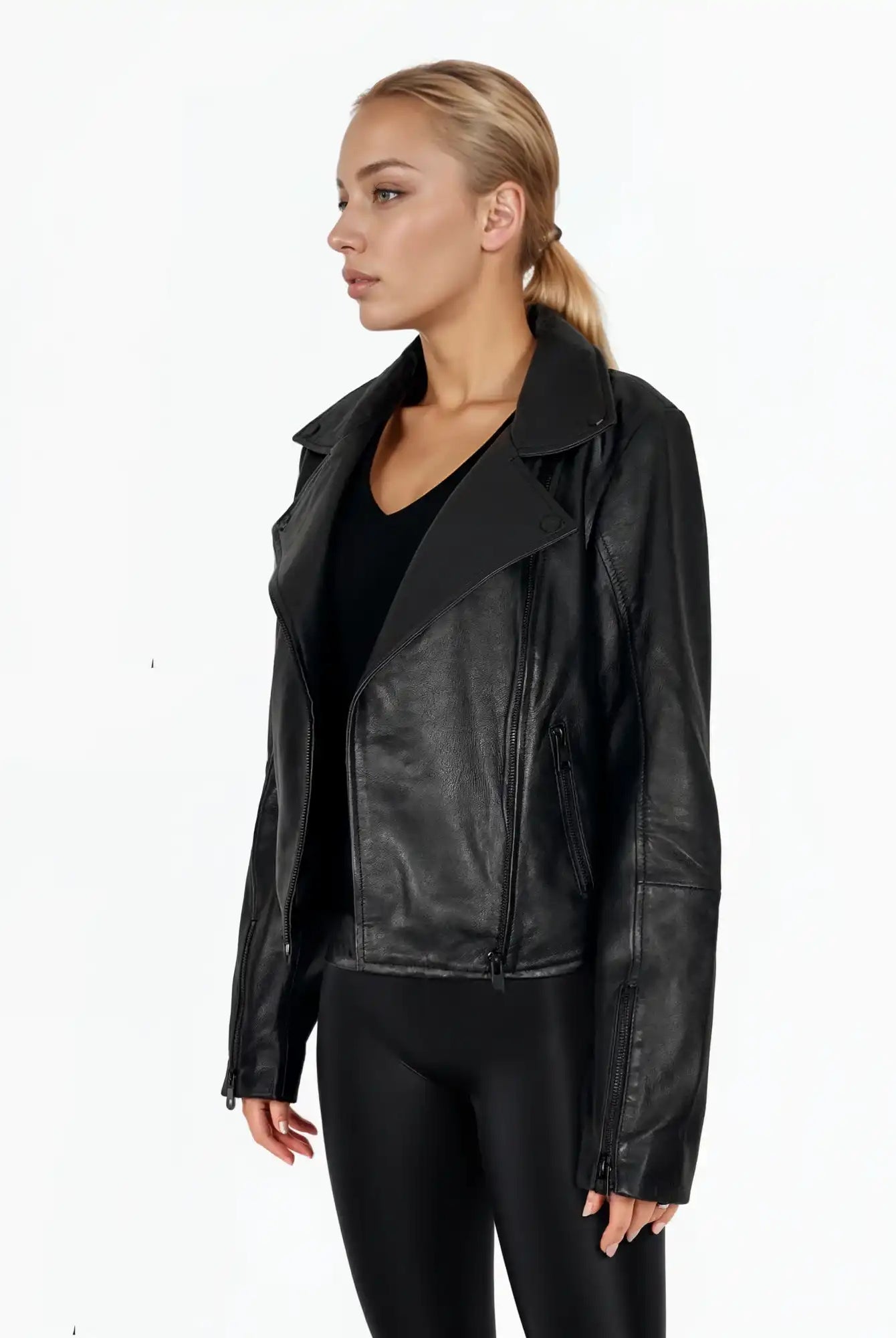 Eine Frau mit zurückgebundenen blonden Haaren trägt die trueprodigy Stella taillierte Lederjacke mit schwarzem Reißverschluss & Ärmelzipper über einem schwarzen Oberteil und Leggings. Sie steht vor einem schlichten weißen Hintergrund und schaut leicht nach links.