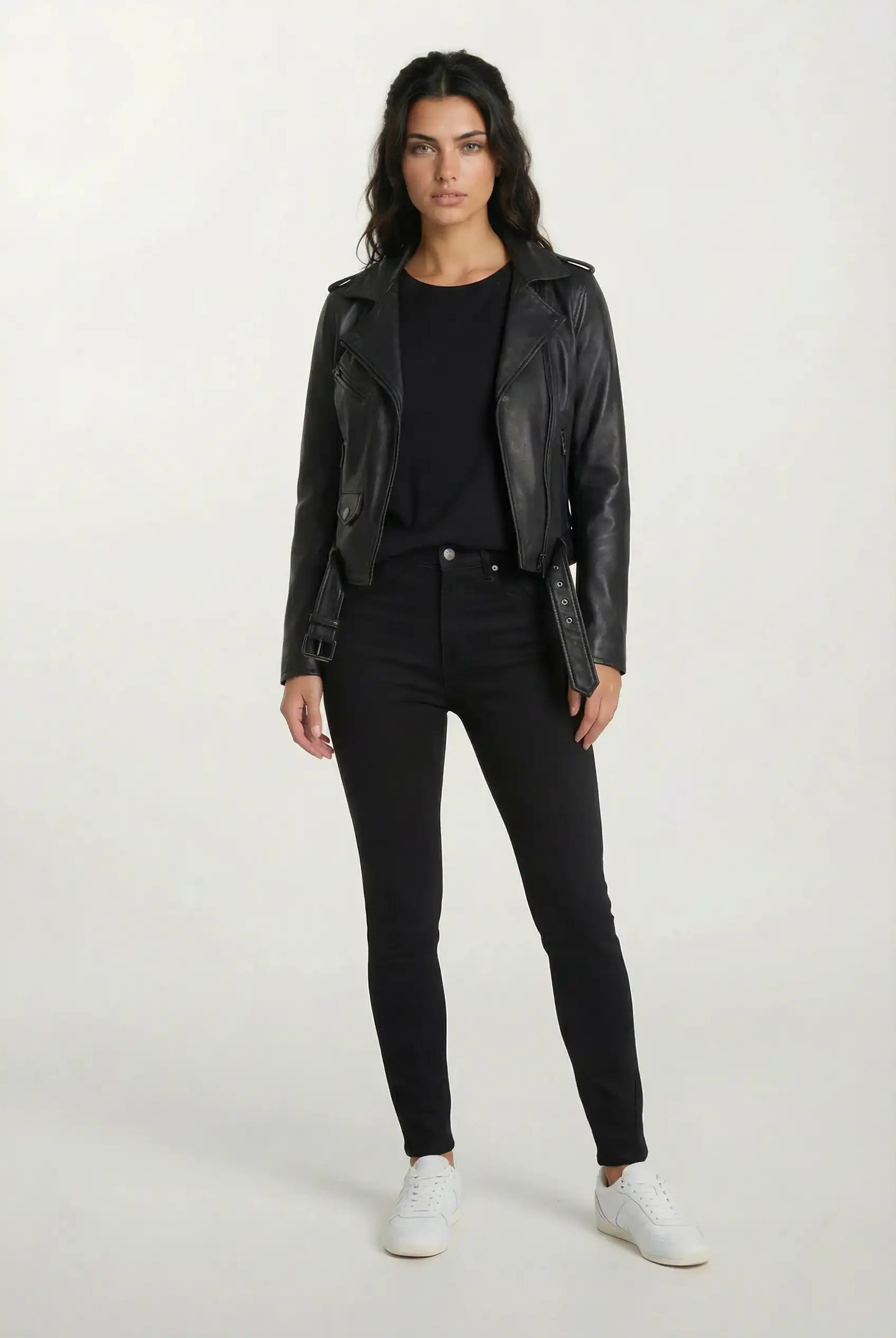 Vor einem hellen Hintergrund trägt eine Frau die trueprodigy Feminine Bikerjacke mit Gürtel & Schulterriegeln, ein schwarzes Top, schwarze Jeans und weiße Sneaker. Sie hat lange dunkle Haare und einen neutralen Gesichtsausdruck.
