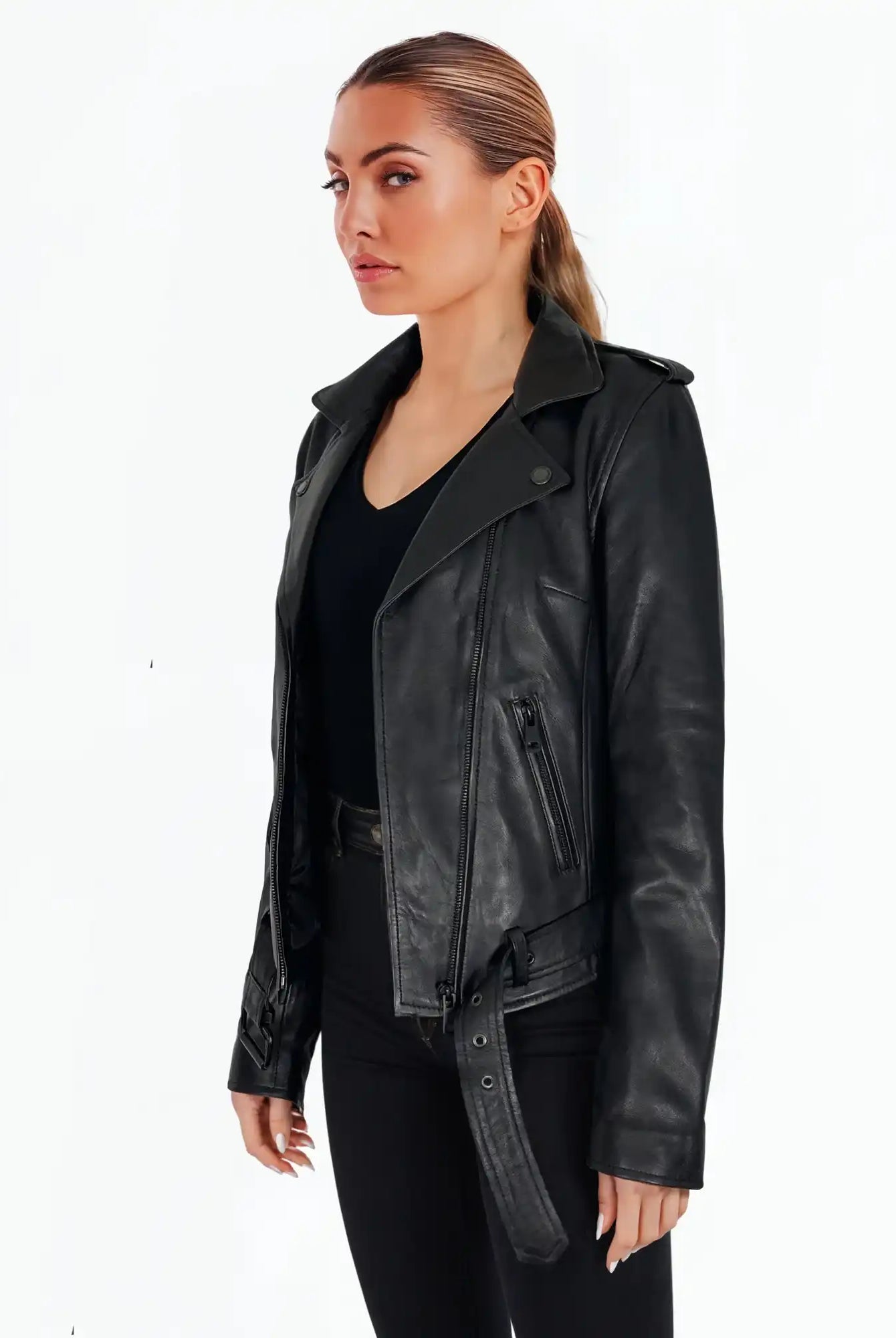 Eine Frau steht vor einem weißen Hintergrund und trägt die trueprodigy Laura Feminine Bikerjacke mit Gürtel & Schulterriegeln über einem schwarzen Oberteil und einer schwarzen Hose. Sie hat ihr Haar zurückgebunden und schaut leicht zur Seite.