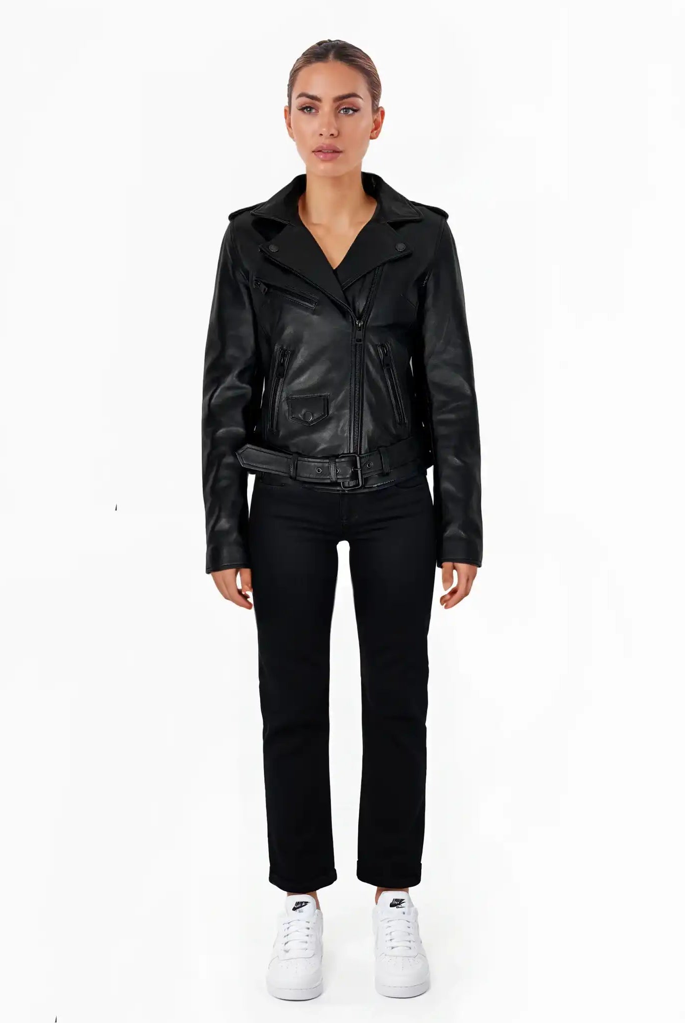 Eine Frau steht mit dem Gesicht nach vorne vor einem weißen Hintergrund und trägt die trueprodigy Laura Feminine Bikerjacke mit Gürtel & Schulterriegeln, eine schwarze Hose und weiße Turnschuhe. Ihre Arme ruhen an ihren Seiten und sie hat einen neutralen Gesichtsausdruck.
