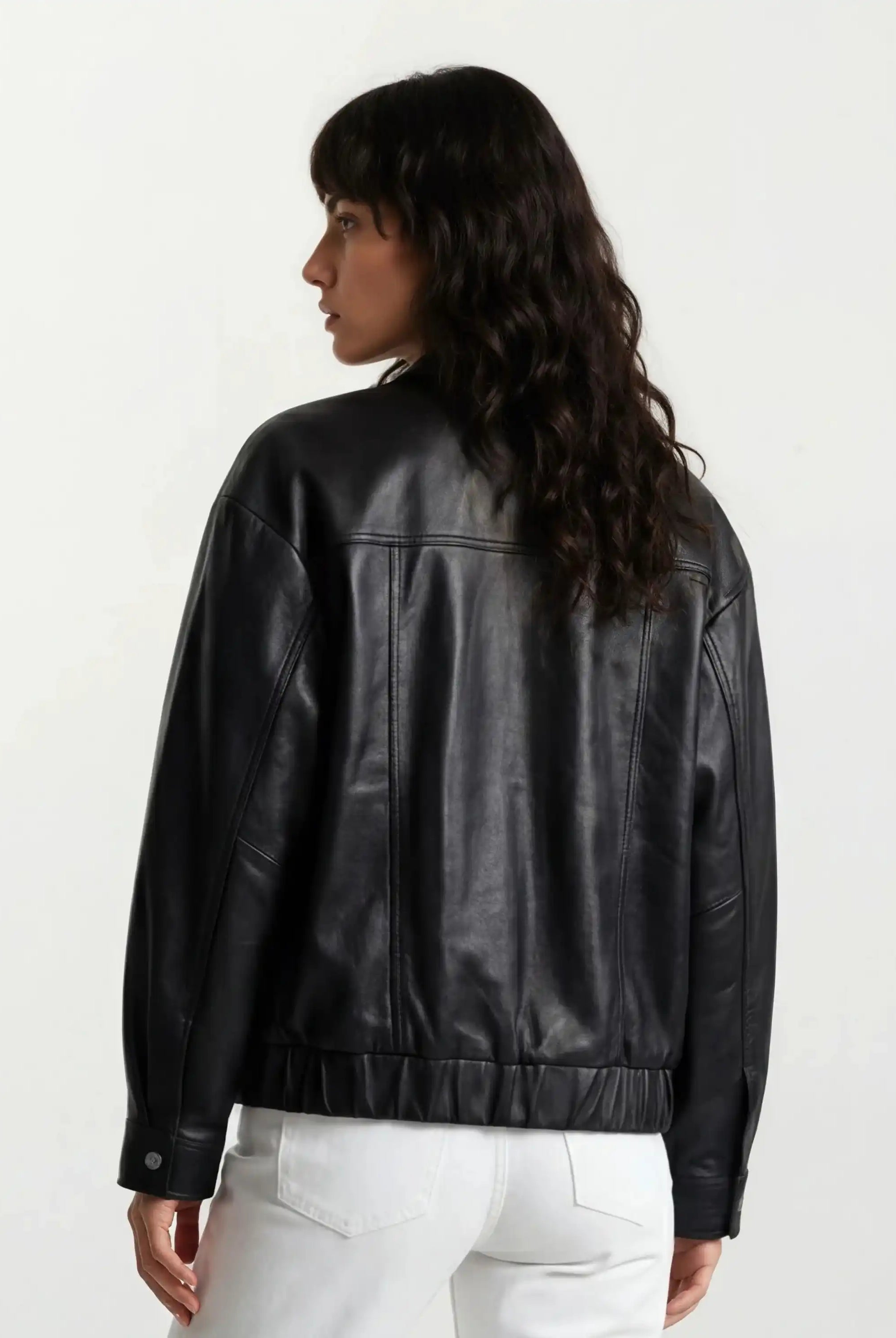 Eine Person mit langen, gewellten dunklen Haaren wird von hinten gezeigt. Sie trägt eine trueprodigy Oversize-Lederjacke mit Gummizugsaum & Lederdetails und eine weiße Hose vor einem schlichten weißen Hintergrund.