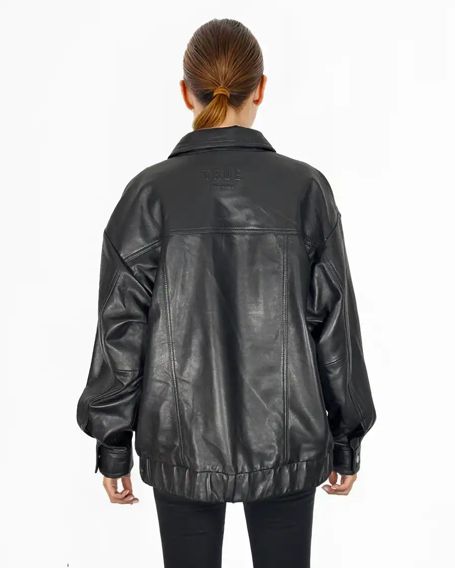 Eine Person mit zurückgebundenen braunen Haaren trägt eine Trueprodigy Nova Oversize-Lederjacke mit Gummizugsaum & Lederdetails und eine schwarze Hose und steht mit dem Rücken zur Kamera vor einem schlichten weißen Hintergrund.