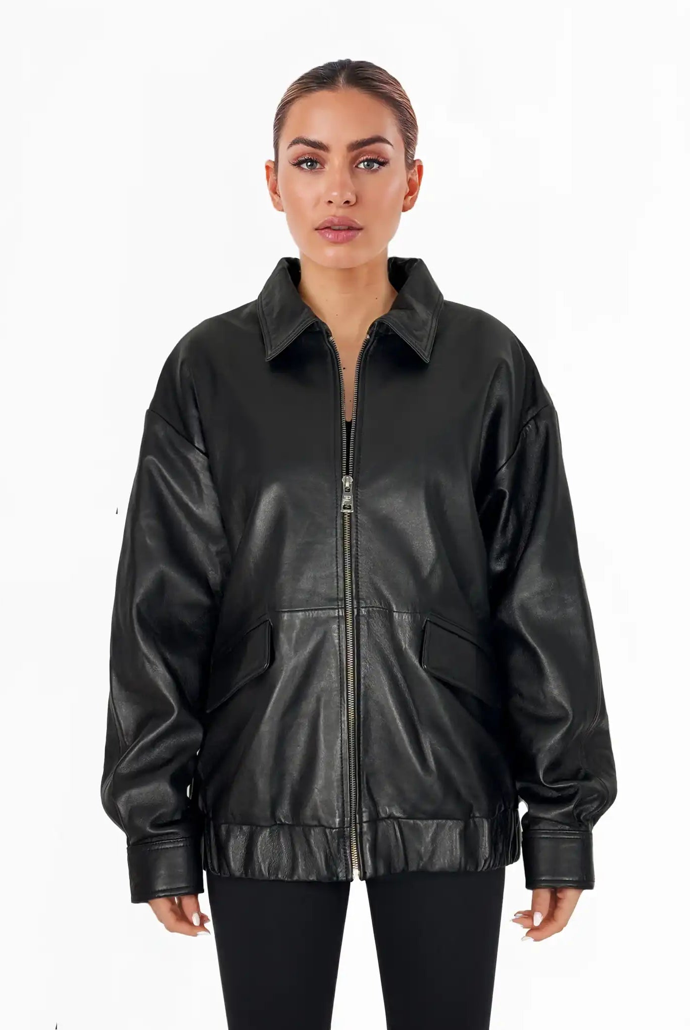 Eine Frau steht mit dem Gesicht nach vorne und trägt die trueprodigy Nova Oversize-Lederjacke mit Gummizugsaum & Lederdetails in Schwarz mit großen Vordertaschen und schwarzen Leggings vor einem schlichten weißen Hintergrund.