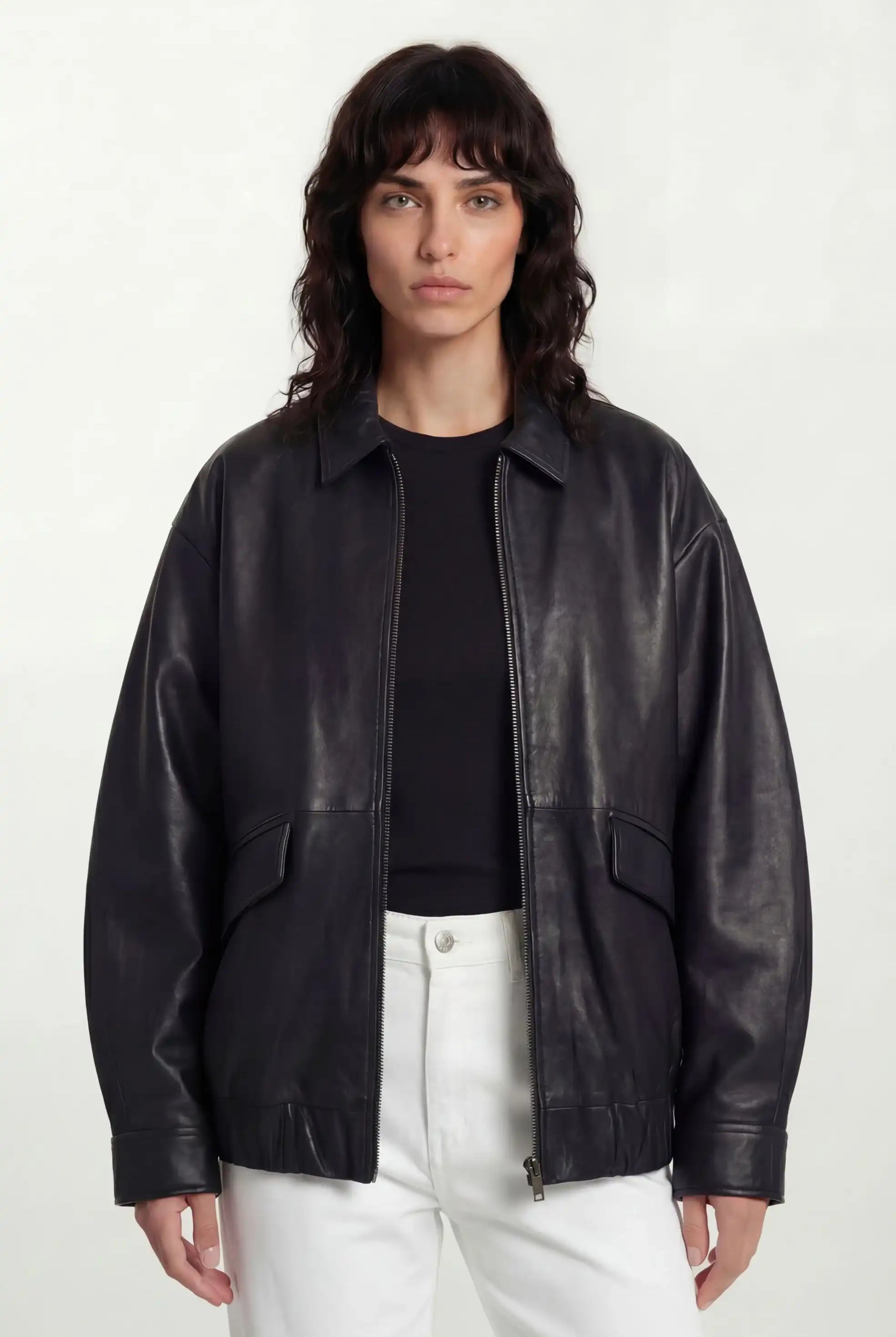 Eine Frau mit gewelltem, schulterlangem dunklem Haar trägt eine trueprodigy Oversize-Lederjacke mit Gummizugsaum & Lederdetails über einem schwarzen Oberteil und einer weißen Hose, posiert vor einem schlichten hellen Hintergrund.