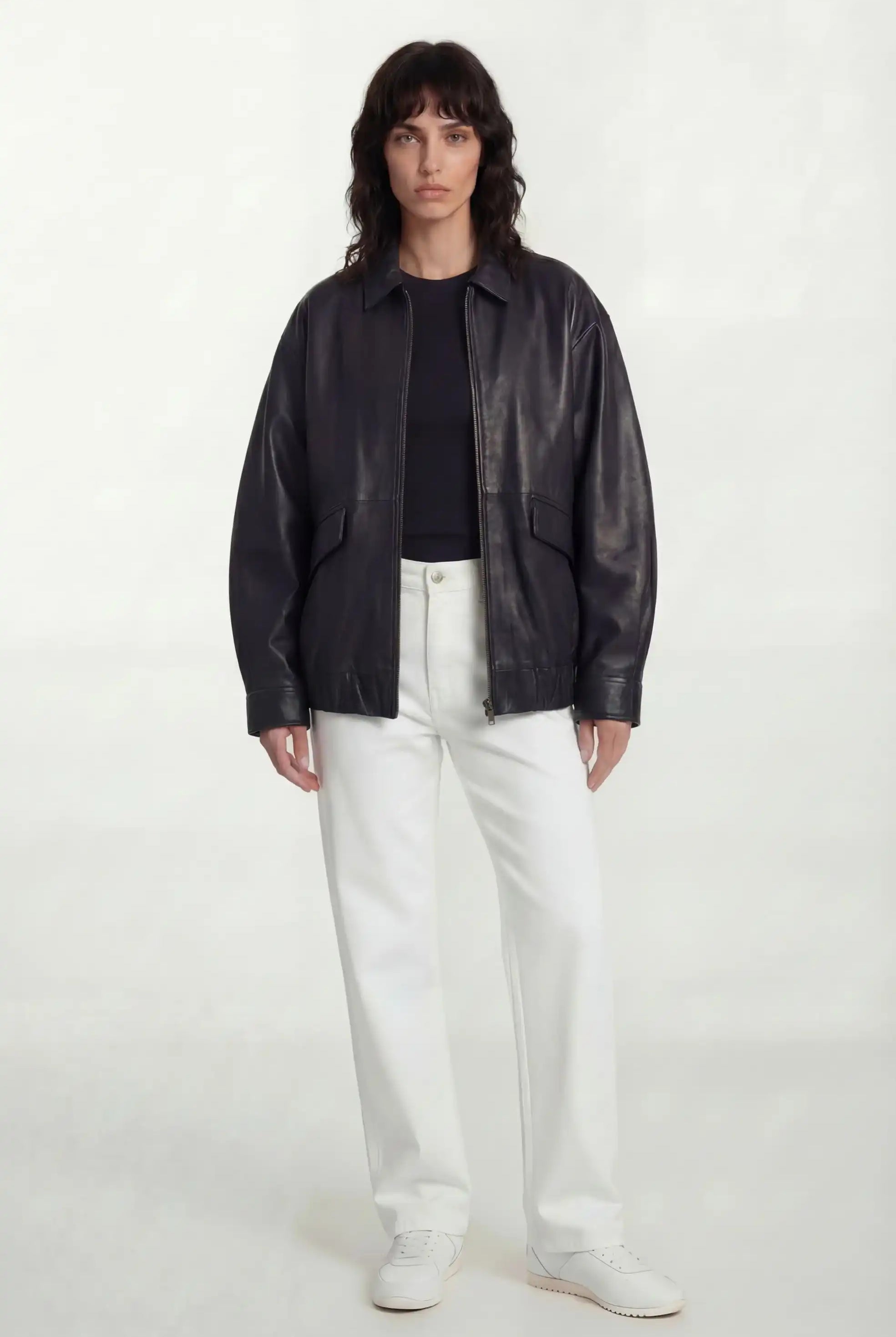 Eine Frau mit mittellangem dunklem Haar trägt die trueprodigy Oversize-Lederjacke mit Gummizugsaum & Lederdetails, ein schwarzes Oberteil, eine weiße Hose und weiße Schuhe. Sie steht vor einem schlichten weißen Hintergrund und blickt mit neutralem Blick nach vorne.