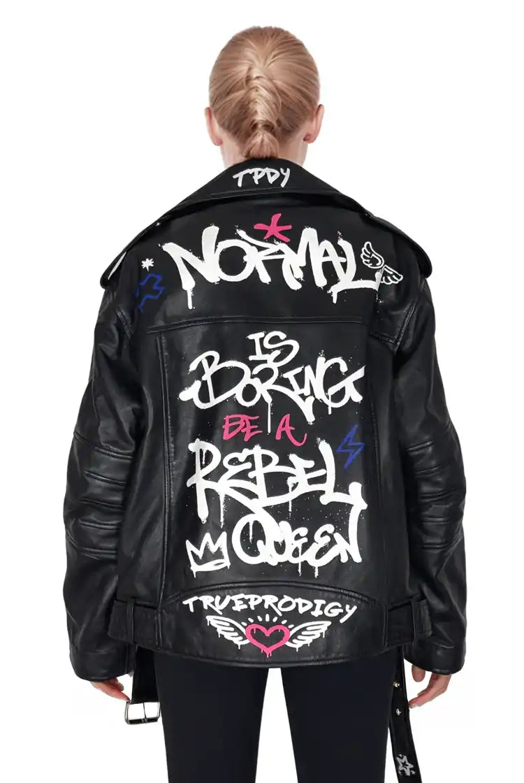 Eine Blondine mit geflochtenen Haaren blickt nach vorne und trägt die trueprodigy "Anya" Schafslederjacke. Auf der Jacke stehen Graffiti-Sprüche wie "Normal ist langweilig", "Sei eine rebellische Königin" und Symbole in Weiß, Pink und Blau - ein mutiges Statement für Frauen.