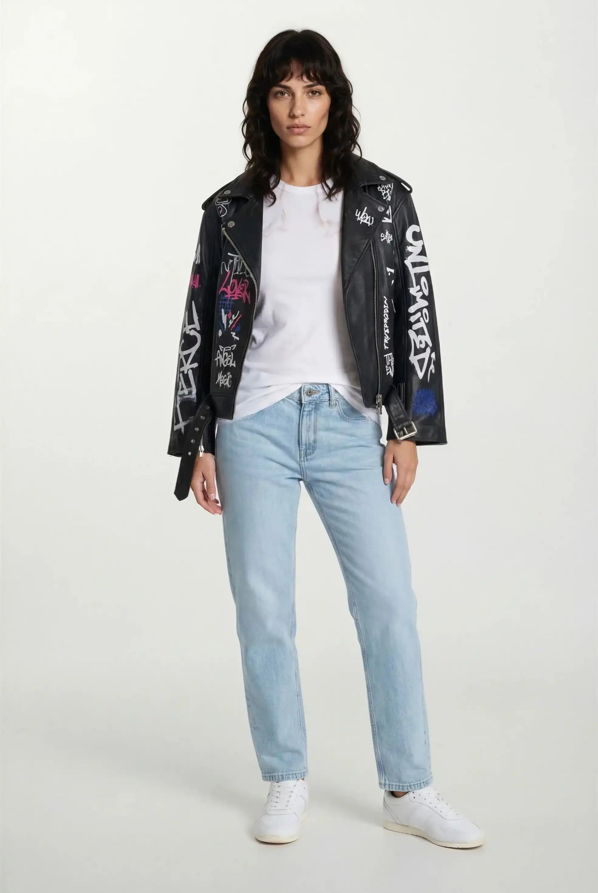 Eine Person in einem weißen T-Shirt, hellblauen Jeans, weißen Turnschuhen und einer trueprodigy Feminine Lederjacke mit Grafittiprint und schrägem Reißverschluss blickt nach vorne. Der Hintergrund ist einfarbig weiß.
