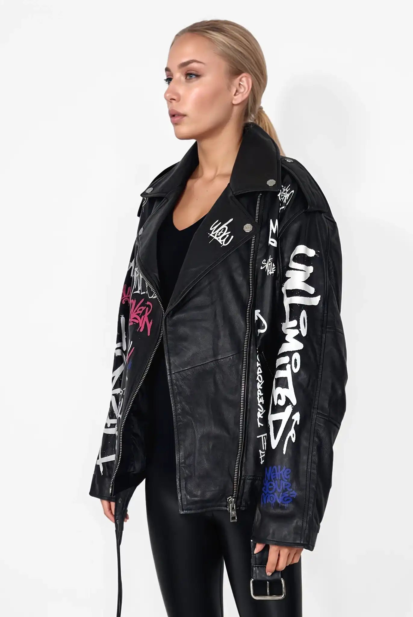 Eine Person mit langen blonden Haaren trägt die schwarze Lederjacke "Anya" von trueprodigy mit weißem und farbigem Graffiti-Text und -Motiven über einem schwarzen Outfit vor einem schlichten weißen Hintergrund.