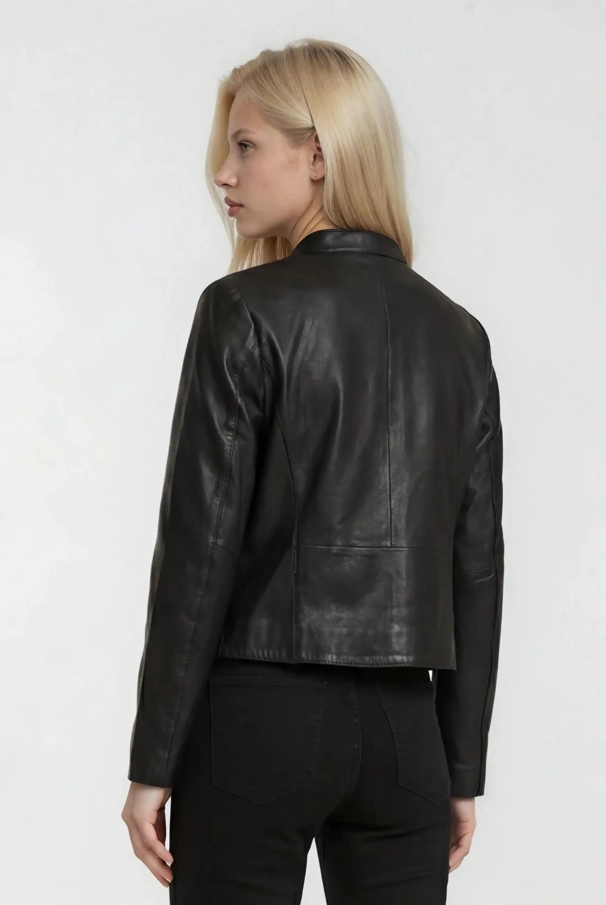 Eine Person mit langen blonden Haaren ist von hinten zu sehen. Sie trägt die trueprodigy Damen Lederjacke aus Schafsleder in Schwarz mit minimalistischem Design, gepaart mit einer schwarzen Lederhose vor einem schlichten hellen Hintergrund.