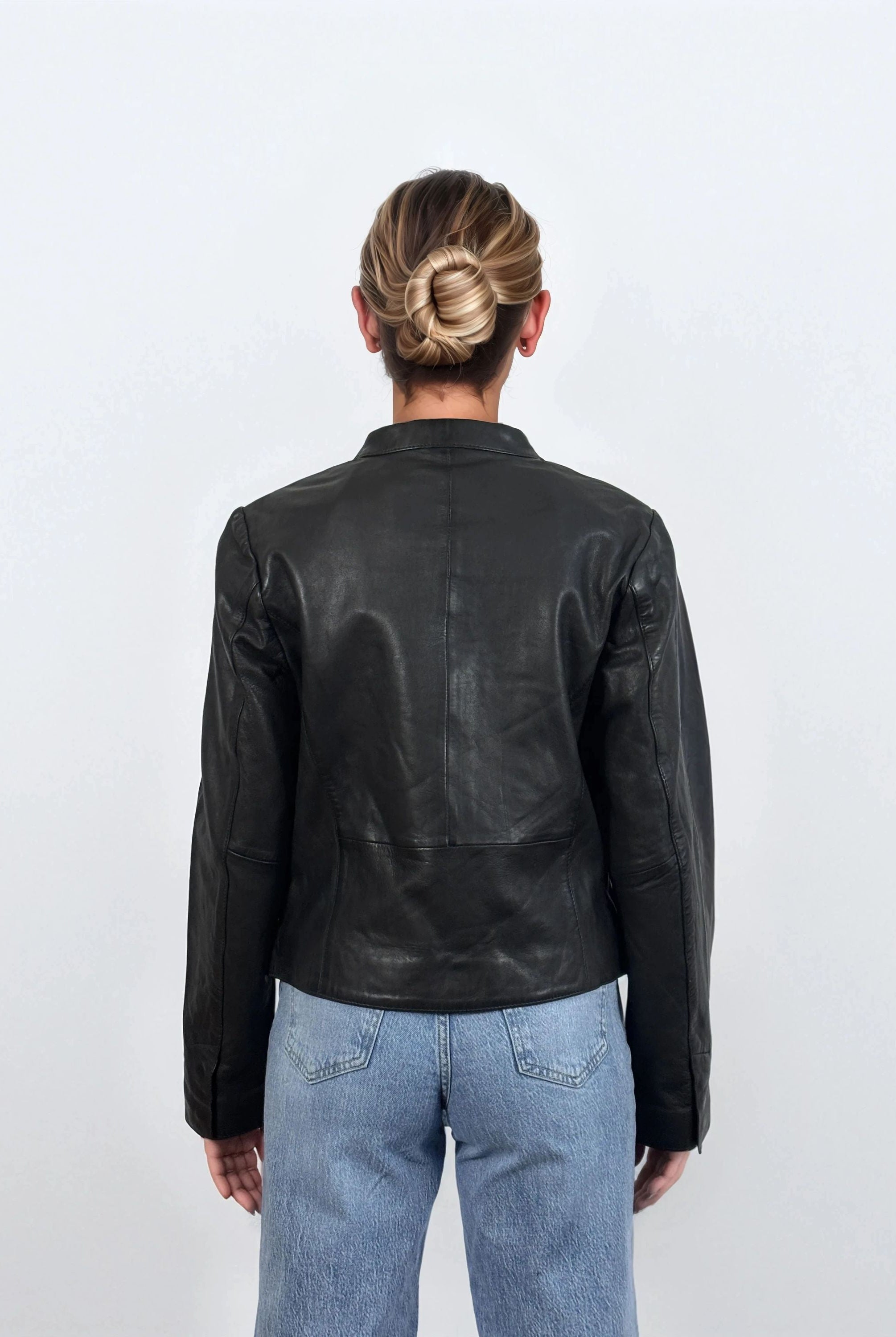 Eine Person mit blondem Haar in einem tiefen Dutt ist von hinten zu sehen. Sie trägt die trueprodigy Damen Lederjacke aus Schafsleder in Schwarz mit minimalistischem Design und hellblaue Jeans vor einem schlichten weißen Hintergrund.
