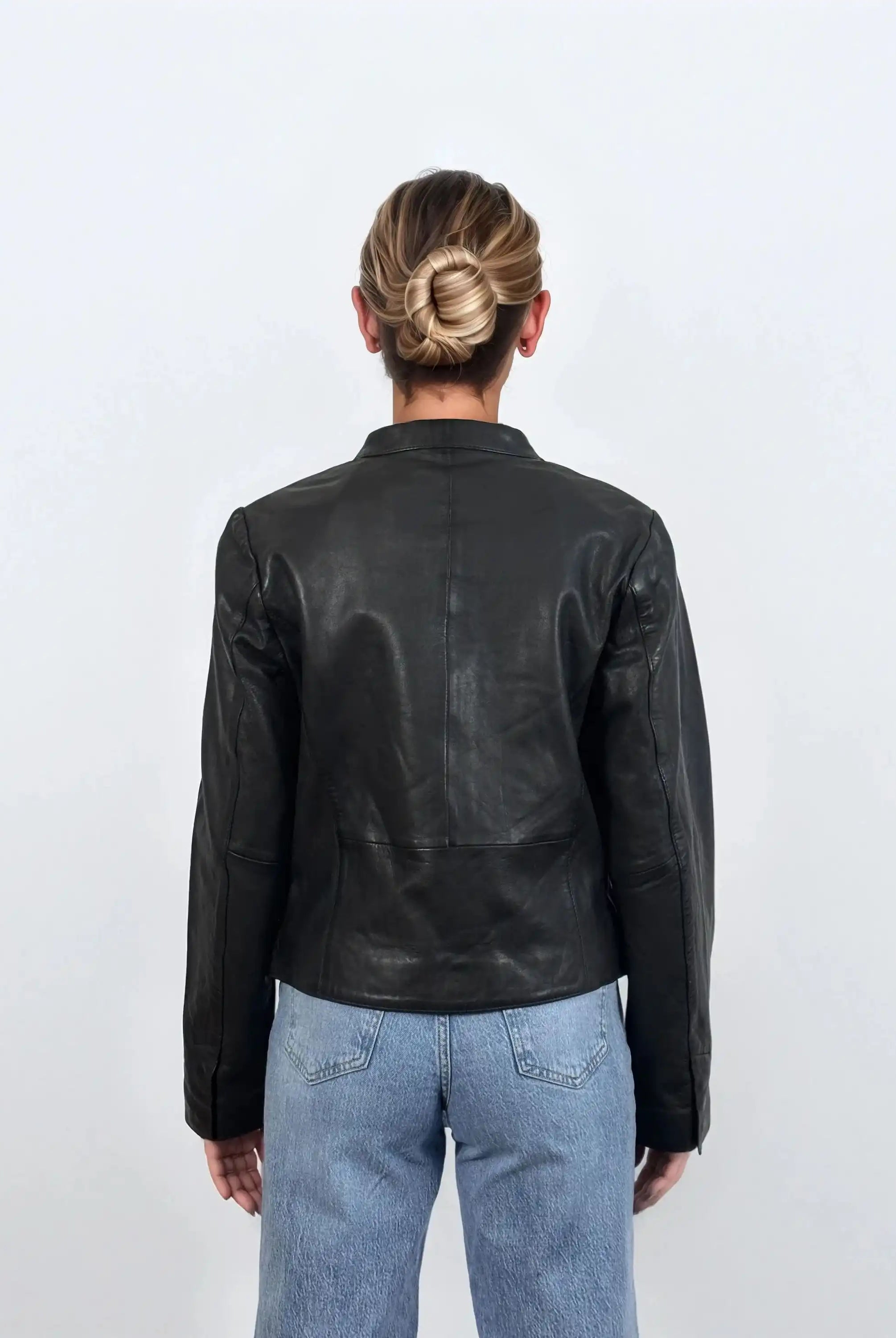 Eine Person mit blondem Haar in einem tiefen Dutt ist von hinten zu sehen. Sie trägt die trueprodigy Damen Lederjacke aus Schafsleder in Schwarz mit minimalistischem Design und hellblaue Jeans vor einem schlichten weißen Hintergrund.