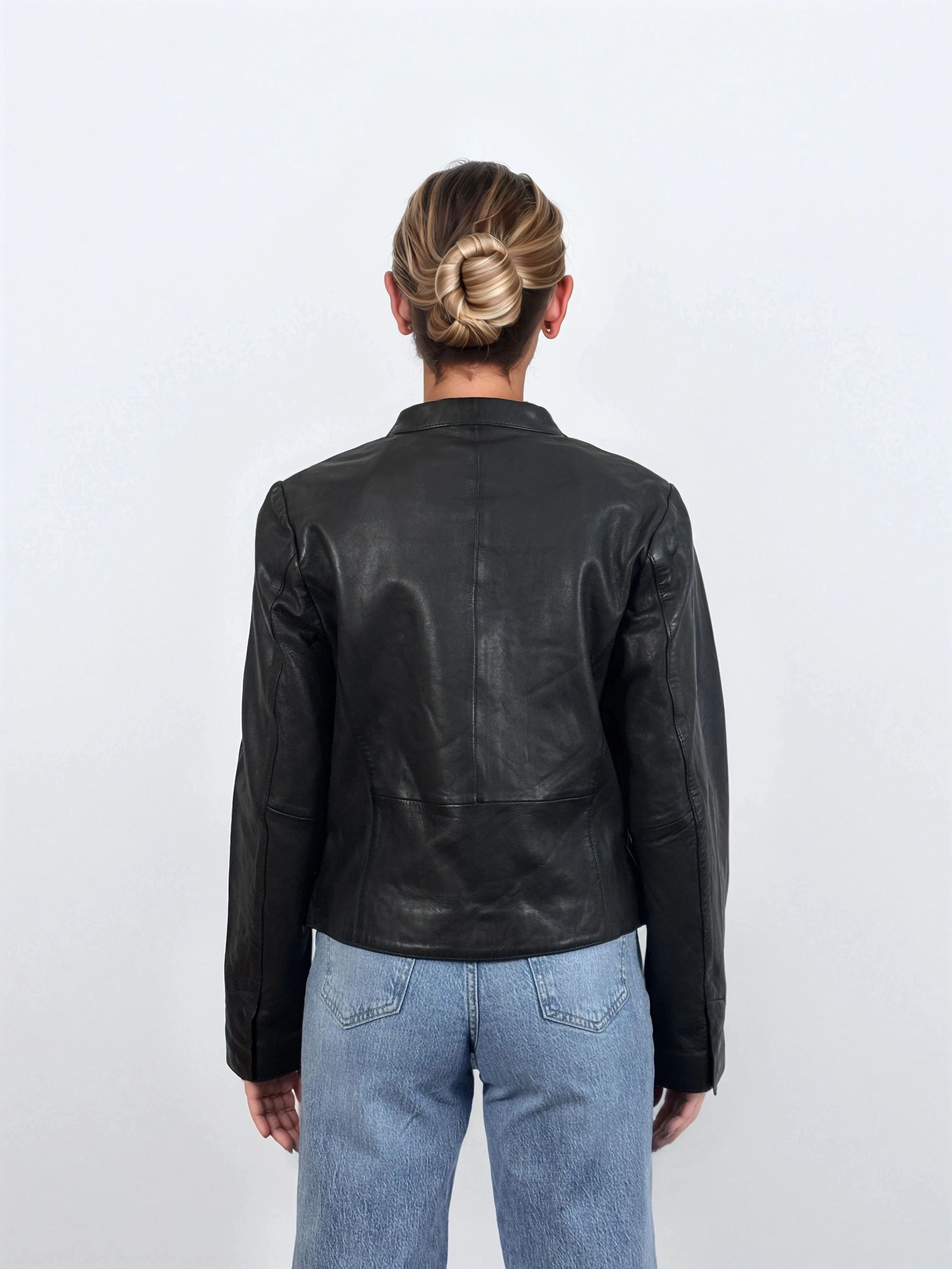 Eine Person mit blondem Haar in einem tiefen Dutt ist von hinten zu sehen. Sie trägt die trueprodigy Damen Lederjacke aus Schafsleder in Schwarz mit minimalistischem Design und hellblaue Jeans vor einem schlichten weißen Hintergrund.