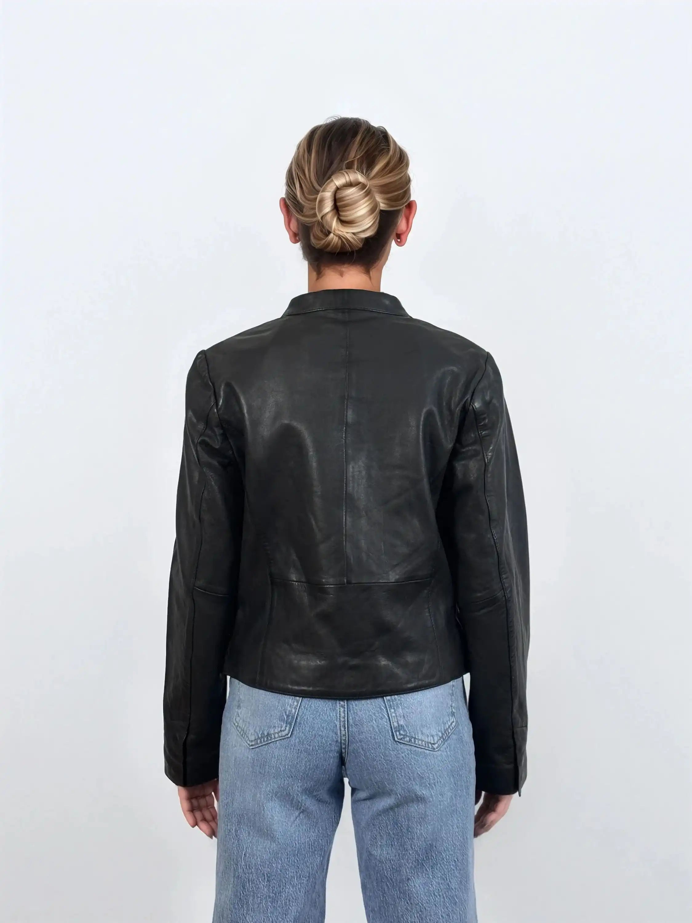 Eine Person mit blondem Haar in einem tiefen Dutt ist von hinten zu sehen. Sie trägt die trueprodigy Damen Lederjacke aus Schafsleder in Schwarz mit minimalistischem Design und hellblaue Jeans vor einem schlichten weißen Hintergrund.