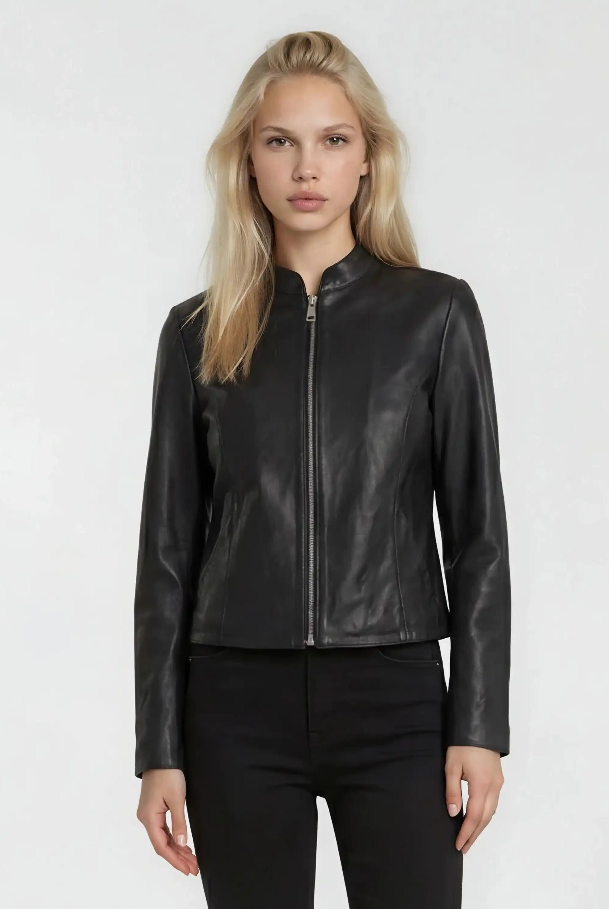 Eine Person mit langen blonden Haaren trägt die trueprodigy Damen Lederjacke aus Schafsleder in Schwarz mit minimalistischem Design und schwarzer Hose, stehend vor einem schlichten hellen Hintergrund.