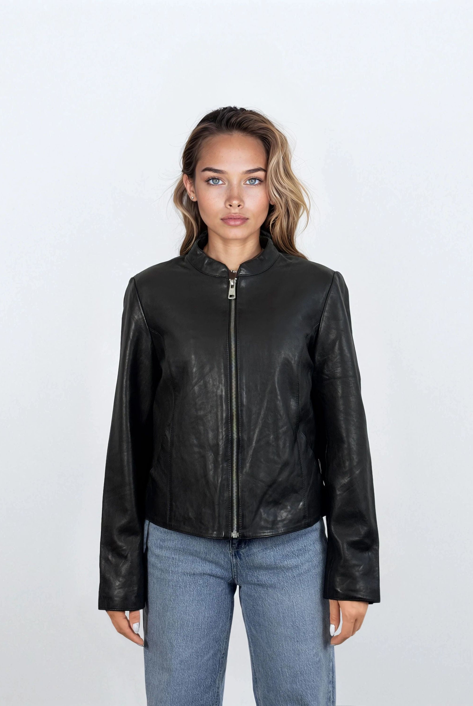 Eine Person mit hellbraunem Haar, die die trueprodigy Damen Lederjacke aus Schafsleder in Schwarz mit minimalistischem Design und Blue Jeans trägt, steht mit dem Gesicht nach vorne vor einem schlichten weißen Hintergrund.