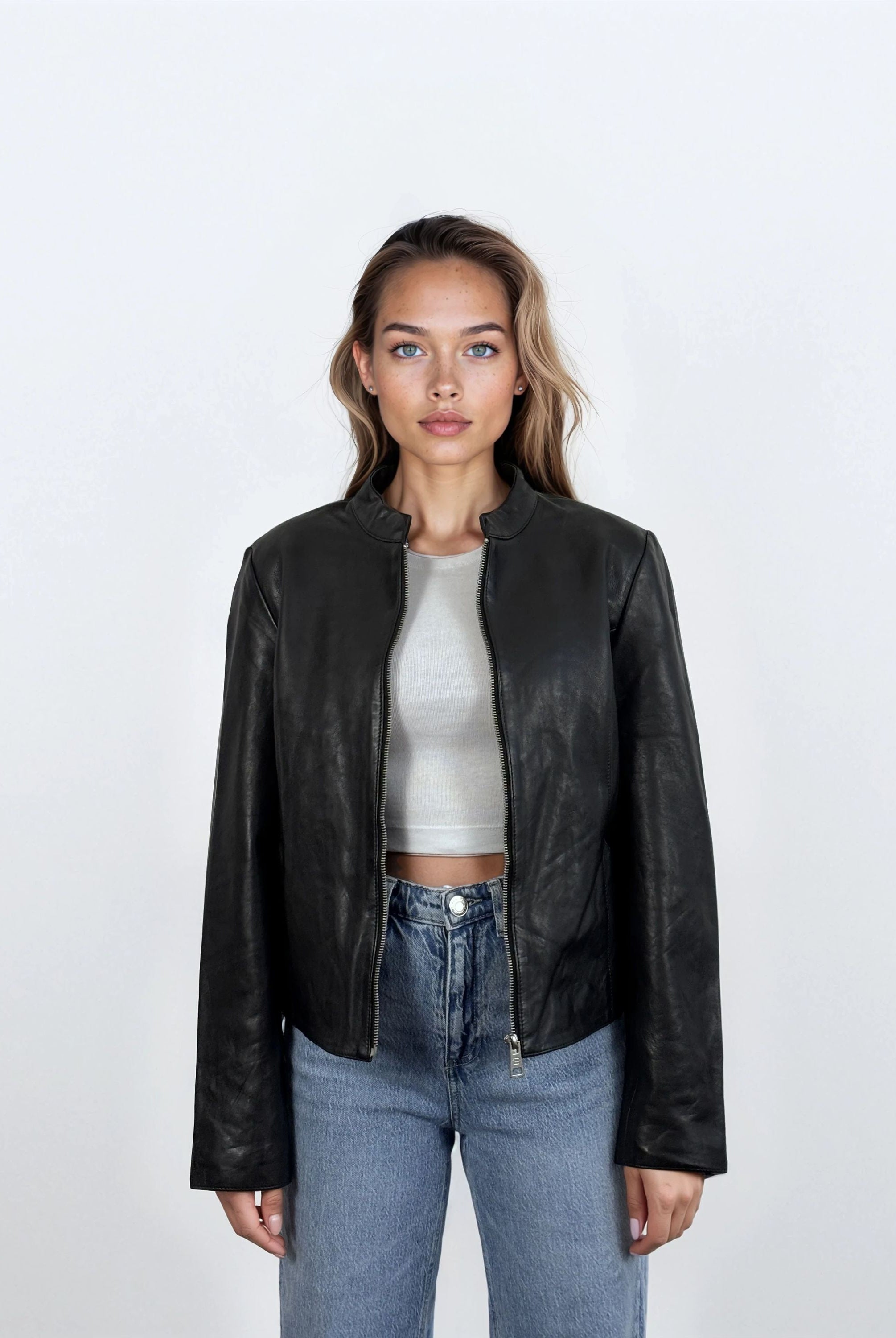 Eine Frau mit langem, gewelltem Haar und neutralem Gesichtsausdruck trägt die trueprodigy Damen Lederjacke aus Schafsleder in Schwarz mit minimalistischem Design über einem hellgrauen Crop-Top und blauen Jeans vor einem schlichten weißen Hintergrund.